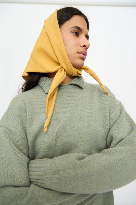 A person stands with arms crossed against a plain white background, wearing a sage green knit sweater and the Extreme Cashmere No. 35 Bandana in Wheat, tied under the chin.