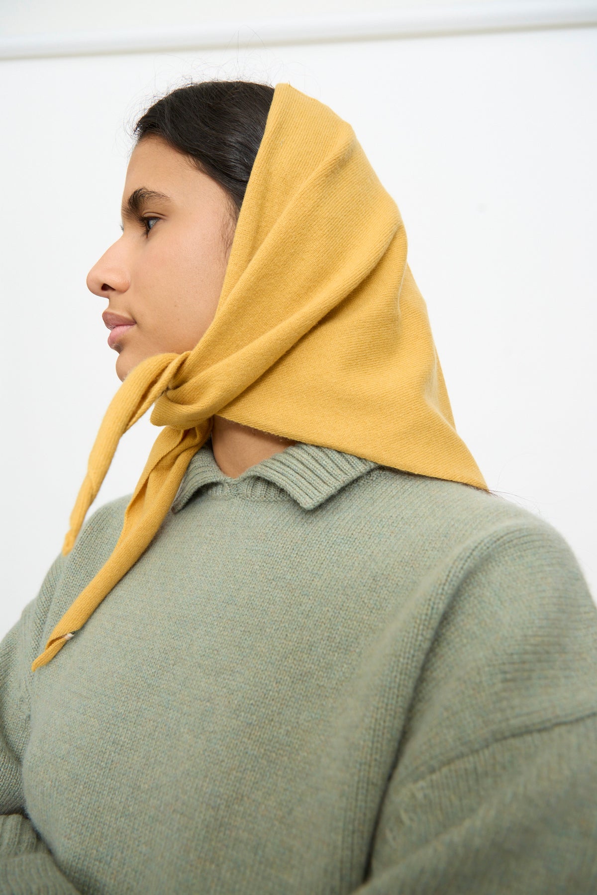 A person in profile wears the Extreme Cashmere No. 35 Bandana in Wheat, tied under the chin, along with a green sweater, posed against a plain white background.