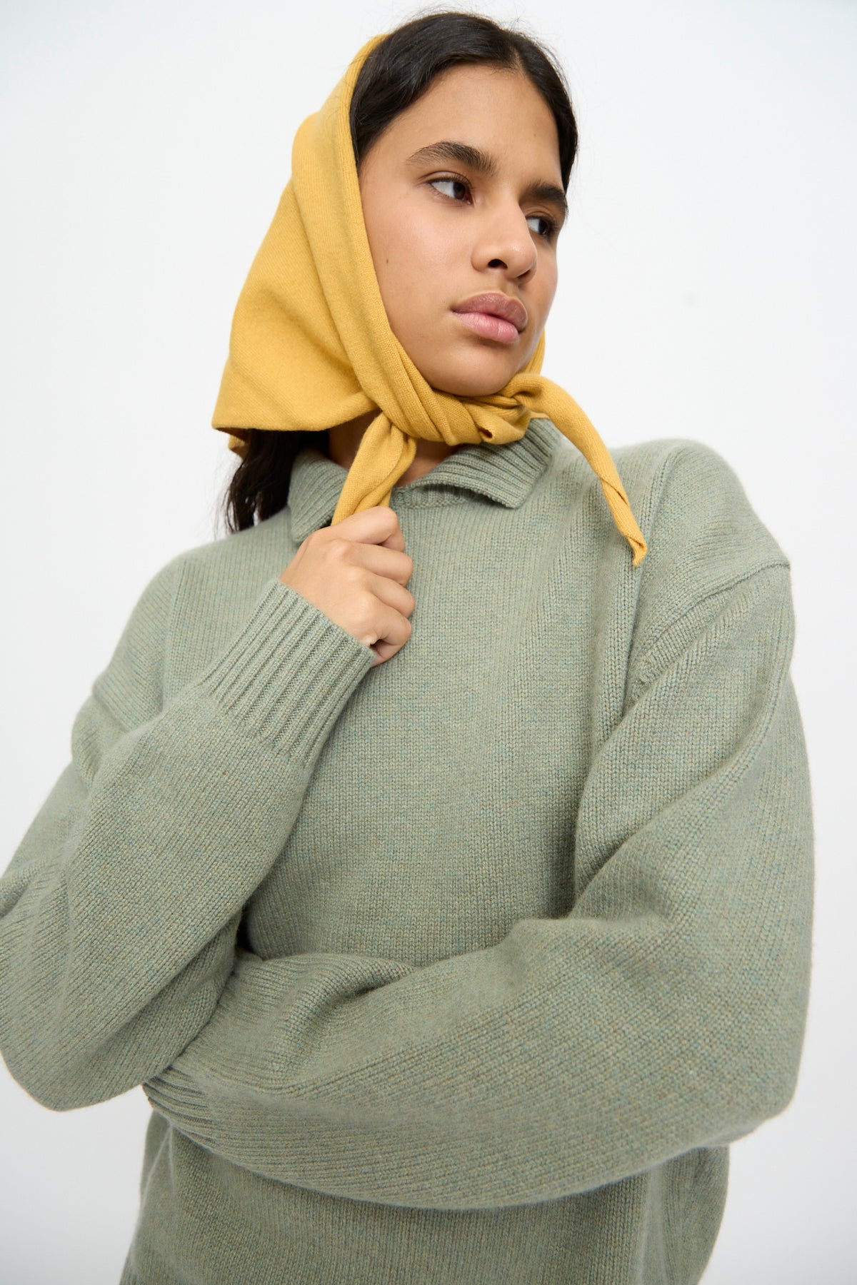 A person wearing a light green sweater and the Extreme Cashmere No. 35 Bandana in Wheat, looking to the side with arms crossed.