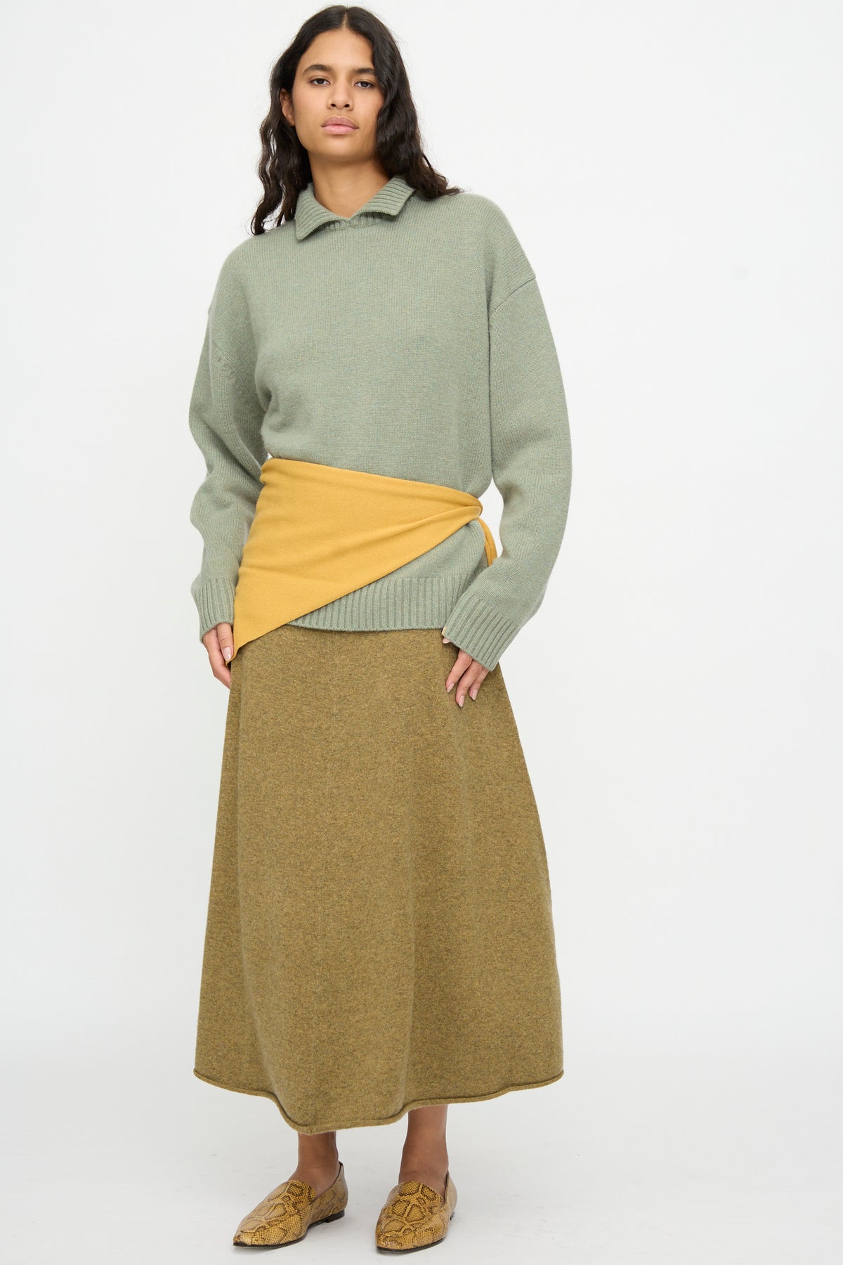 A woman stands against a white background wearing a green sweater, a mustard wrap, a brown skirt, and tan shoes with a geometric pattern. She completes her look with the No. 35 Bandana in Wheat from Extreme Cashmere for an added touch of texture.