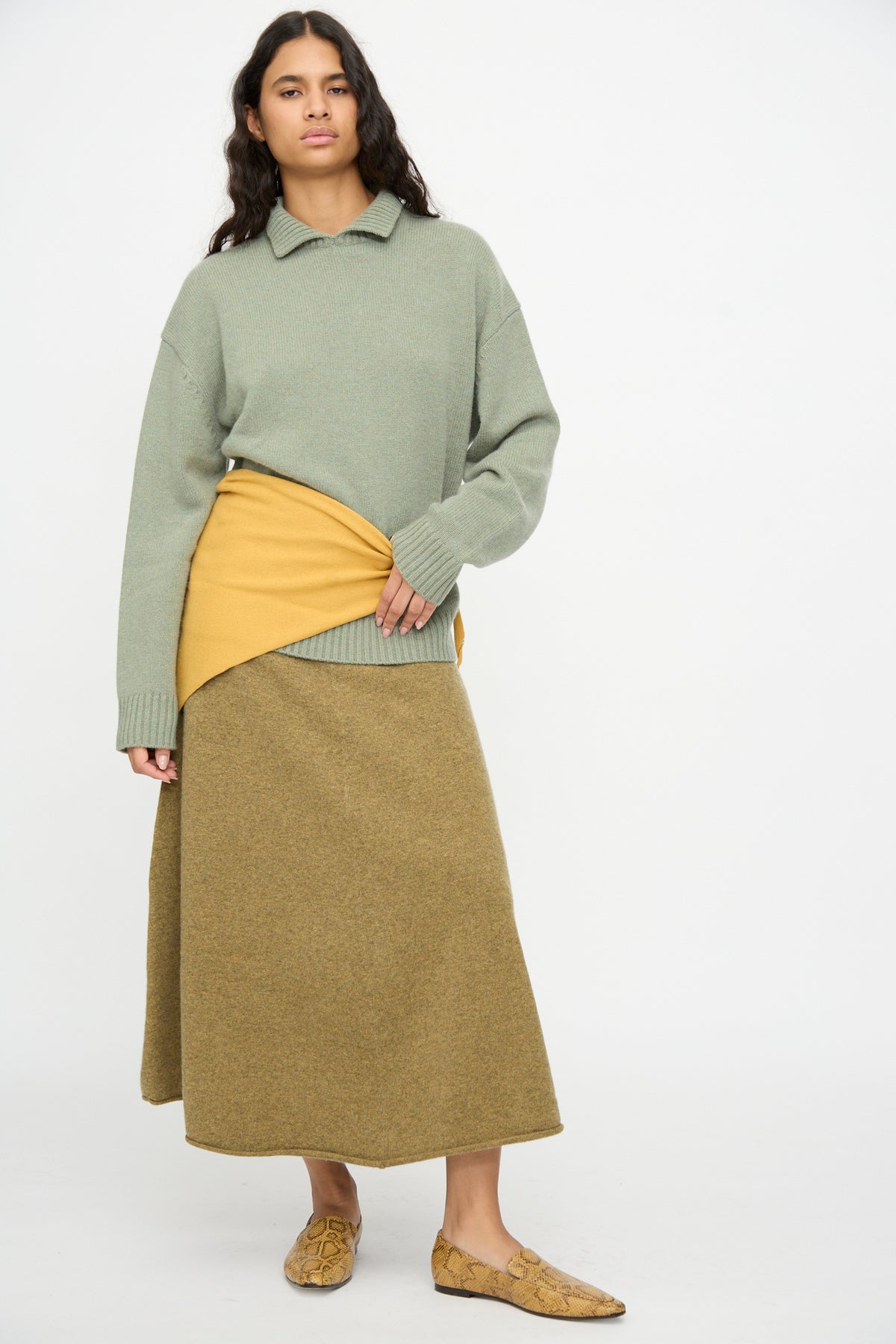 A woman stands against a plain white background, dressed in a green sweater, mustard yellow wrap, olive skirt, and snakeskin-patterned loafers; her look is completed by the Extreme Cashmere No. 35 Bandana in Wheat, elegantly draped at her neck.