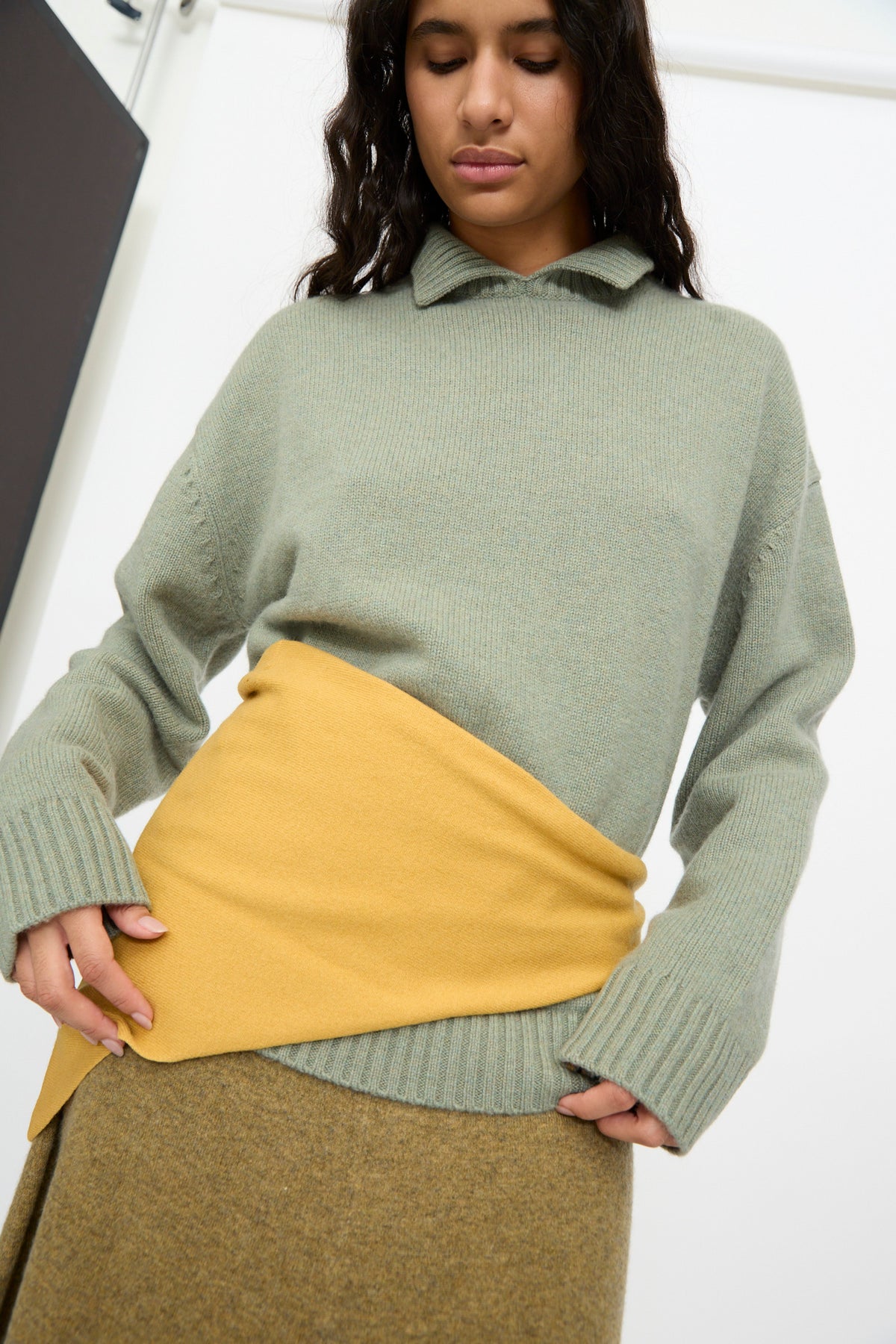 Person wearing a light green sweater with a yellow wrap around the waist, over a brown skirt, standing indoors against a white background, accessorized with the Extreme Cashmere No. 35 Bandana in Wheat.