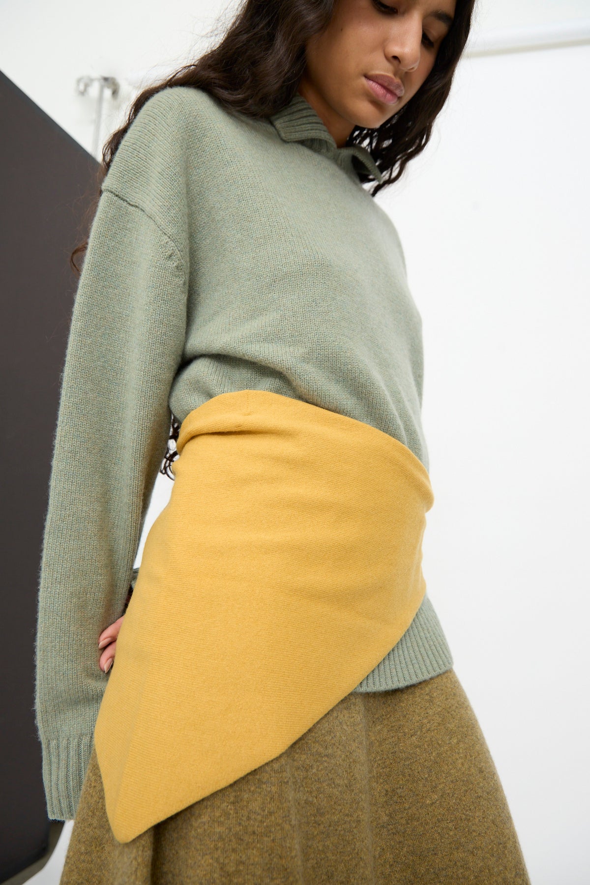 A person wearing a sage green sweater, a mustard yellow asymmetrical wrap skirt, and the No. 35 Bandana in Wheat by Extreme Cashmere stands indoors against a plain background.