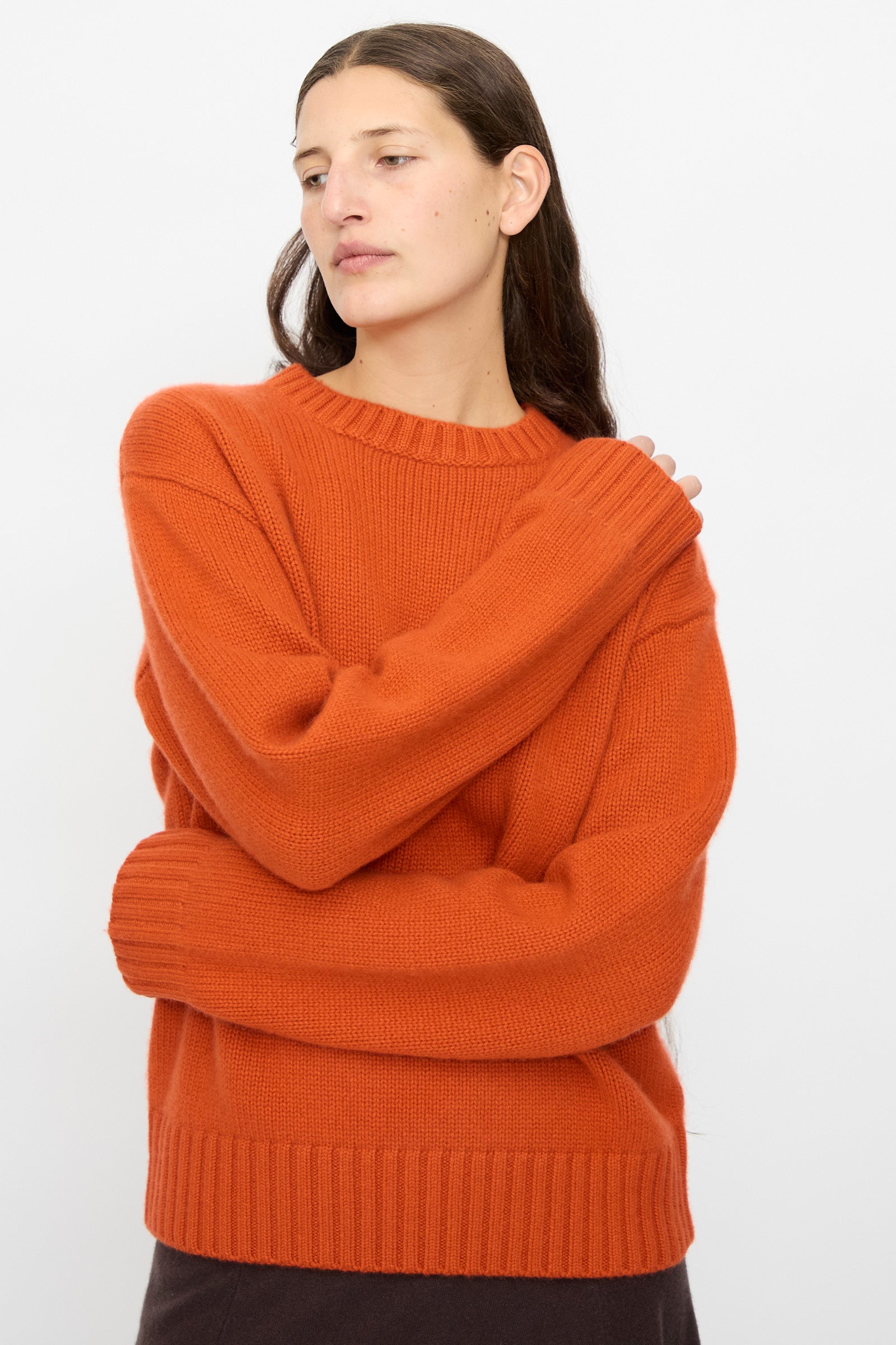 A person with long brown hair wears the Extreme Cashmere No. 373 Oldie Sweater in Dark Orange and dark pants, standing against a plain white background.