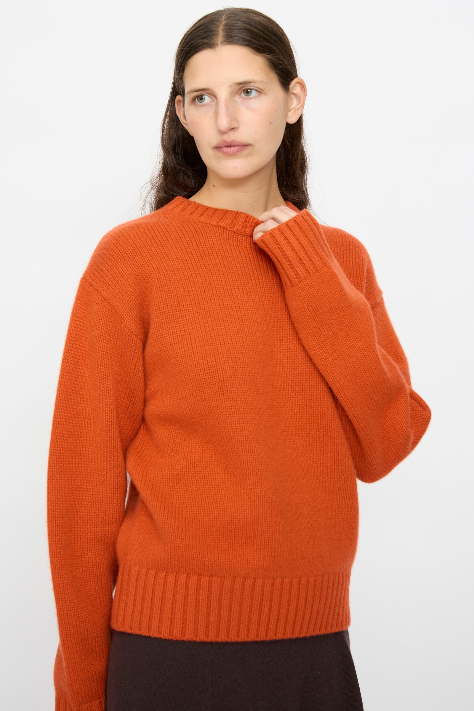 A person with long brown hair wears the Extreme Cashmere No. 373 Oldie Sweater in Dark Orange and dark pants, standing against a plain white background.