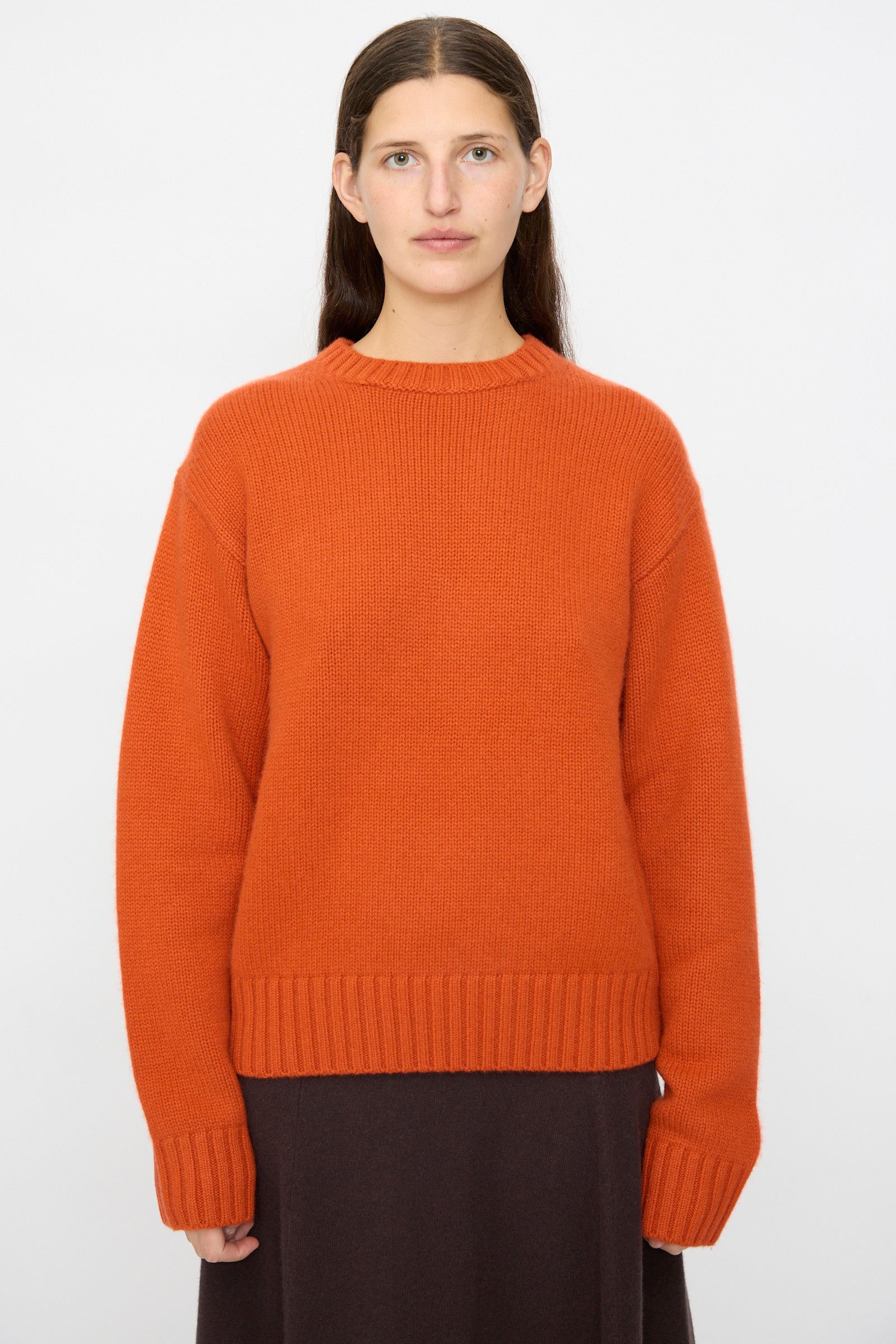 A woman with long brown hair wears the Extreme Cashmere No. 373 Oldie Sweater in Dark Orange and a dark brown skirt, standing against a plain white background.