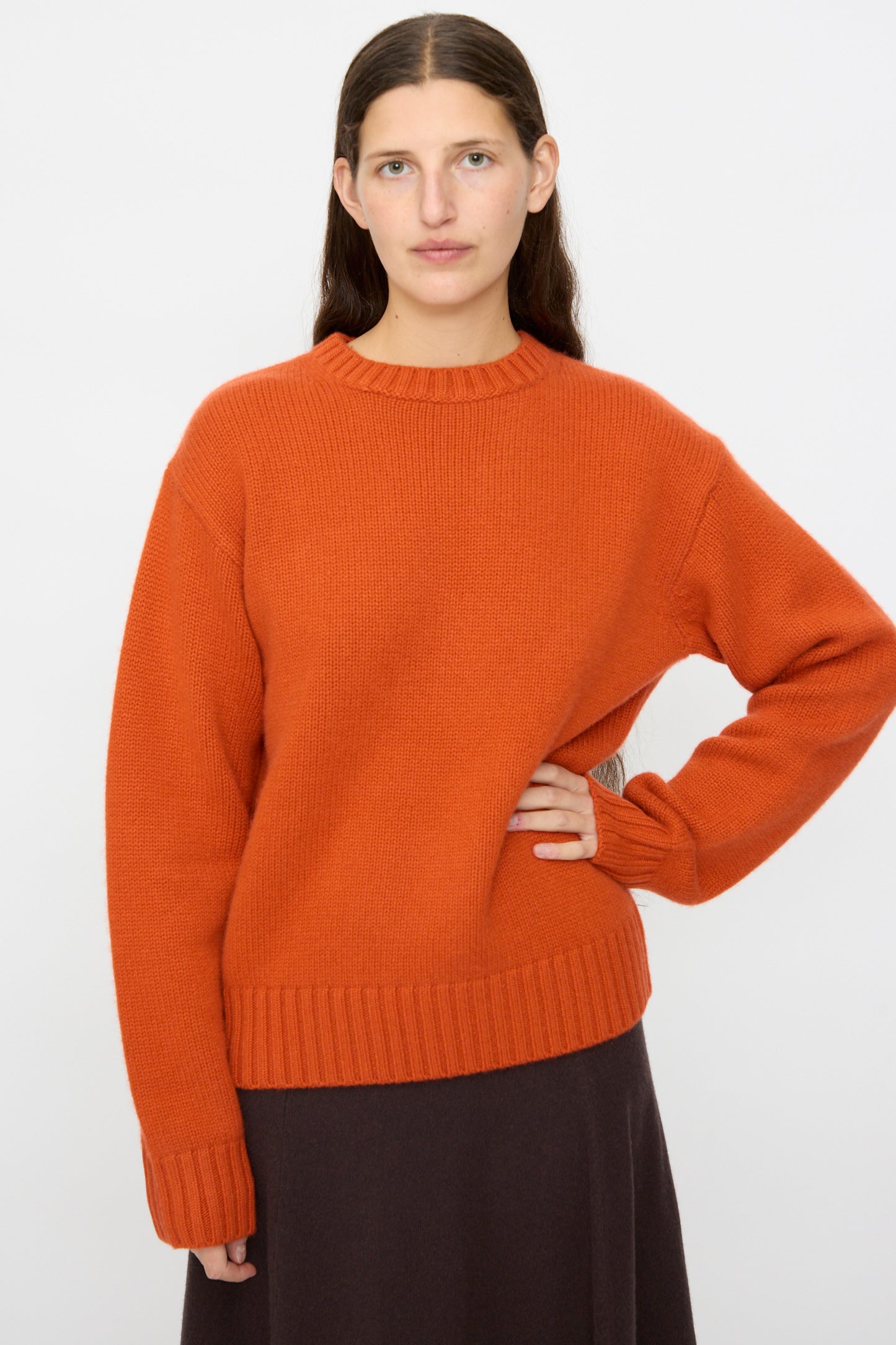 A person with long brown hair wears the Extreme Cashmere No. 373 Oldie Sweater in Dark Orange, relaxed fit, paired with a dark skirt, standing against a plain white background.
