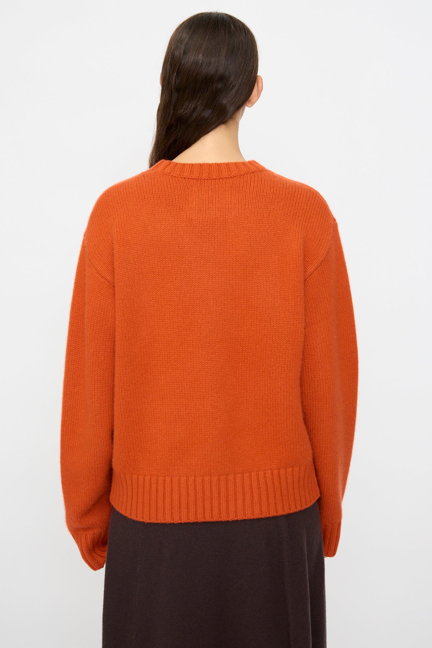 Person with long dark hair wears the Extreme Cashmere No. 373 Oldie Sweater in Dark Orange, styled with a dark skirt and photographed from the back against a plain light background.
