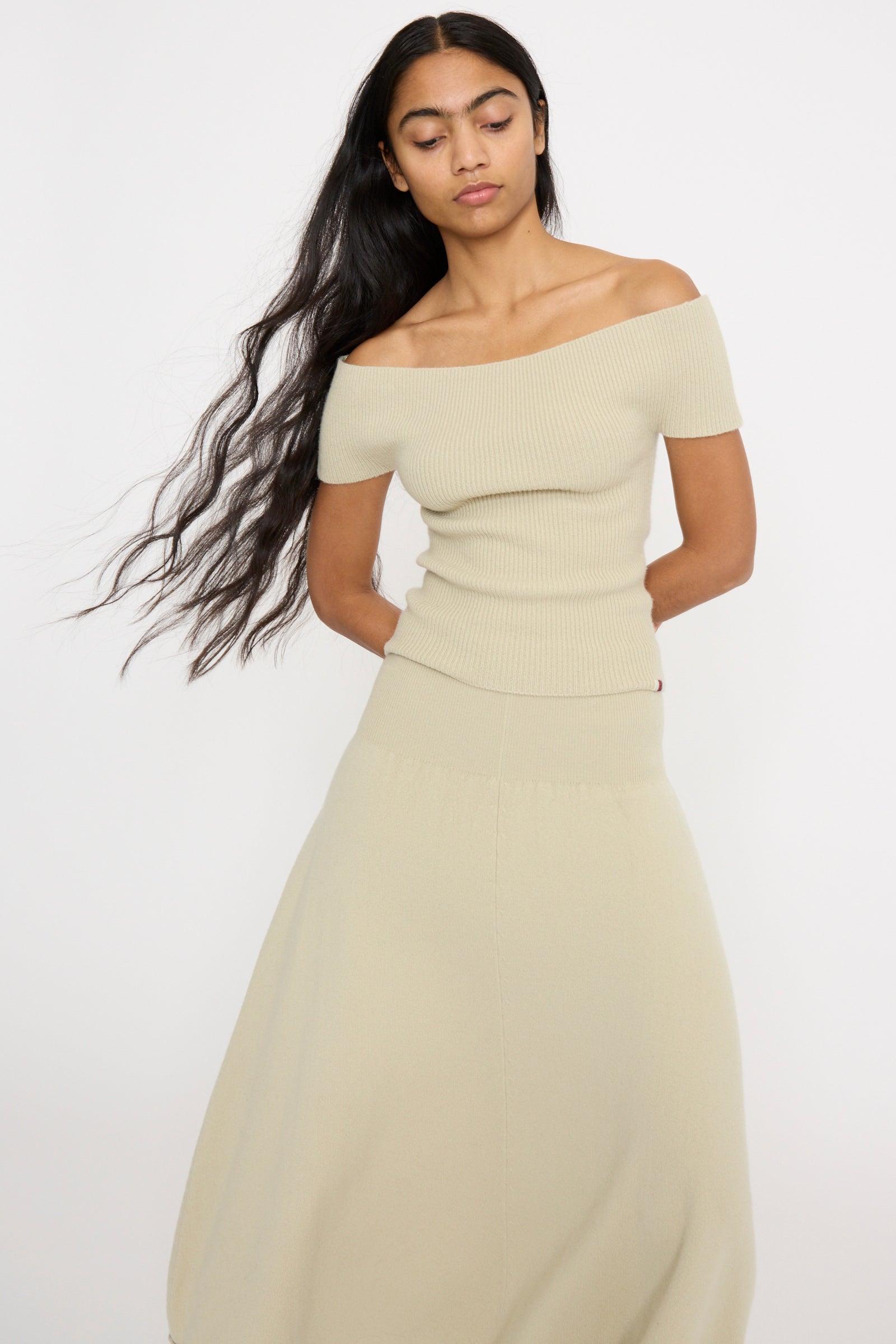 Against a plain white backdrop, a person with long hair wears the Extreme Cashmere No. 381 Libra Sweater in Desert, a slim-fit off-the-shoulder cashmere rib knit top matched with a long skirt.