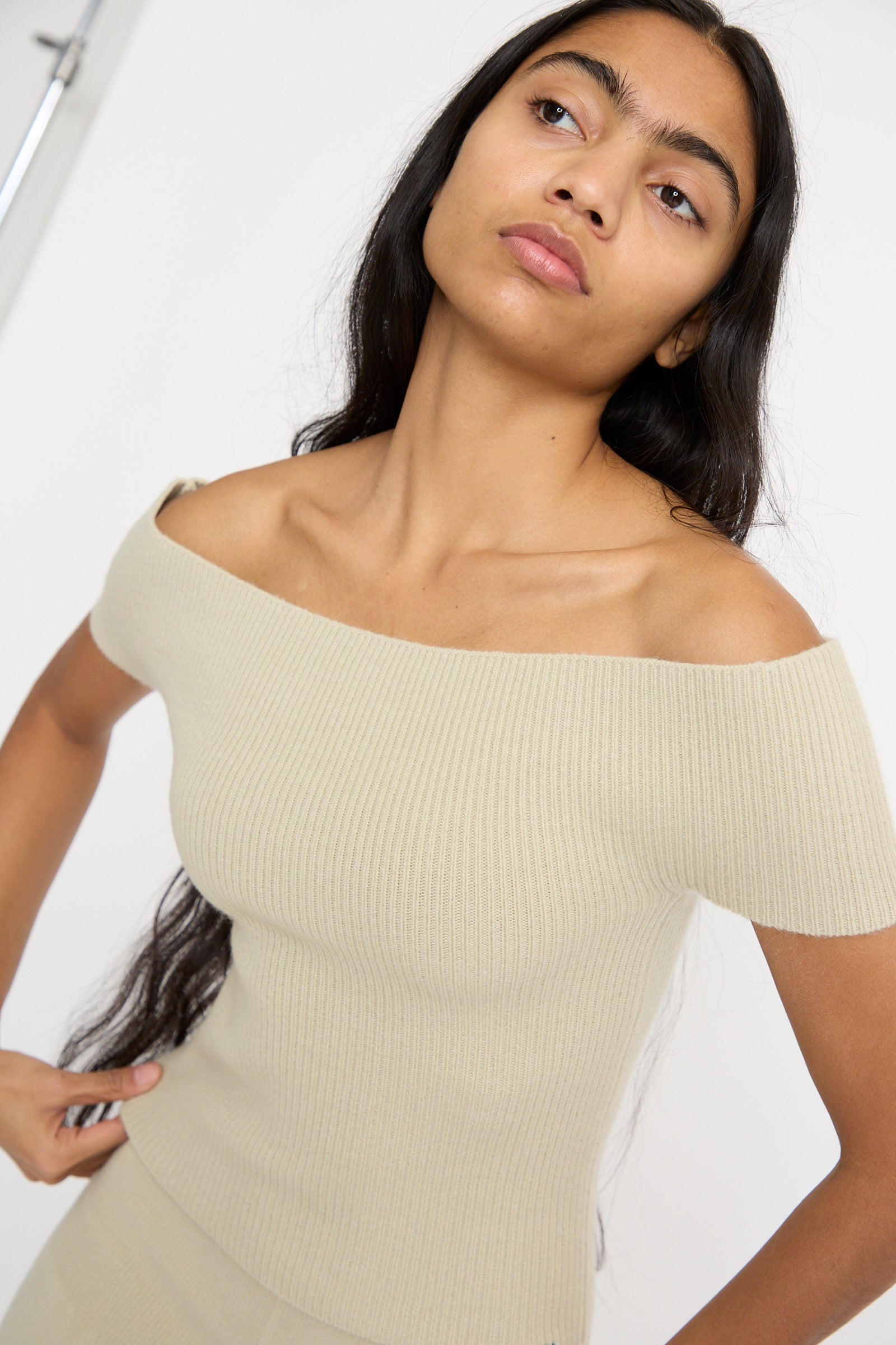 A model wearing the Extreme Cashmere No. 381 Libra Sweater in Desert, a beige cashmere rib knit off-the-shoulder, stands against a neutral background. 