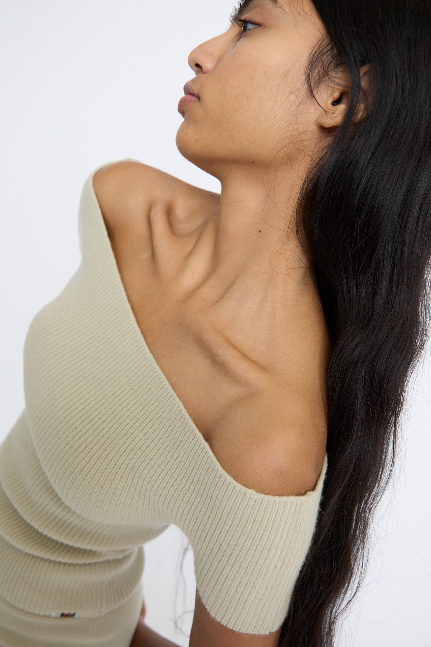 A person with long, dark hair poses in profile against a plain background, wearing the slim-fit off-shoulder No. 381 Libra Sweater in Desert by Extreme Cashmere, crafted from light-colored cashmere rib knit.