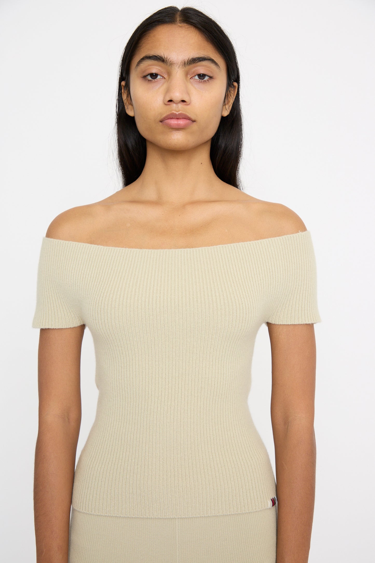 A person with long dark hair stands against a plain white background, wearing the Extreme Cashmere No. 381 Libra Sweater in Desert, a slim-fit off-the-shoulder top made from beige cashmere rib knit.