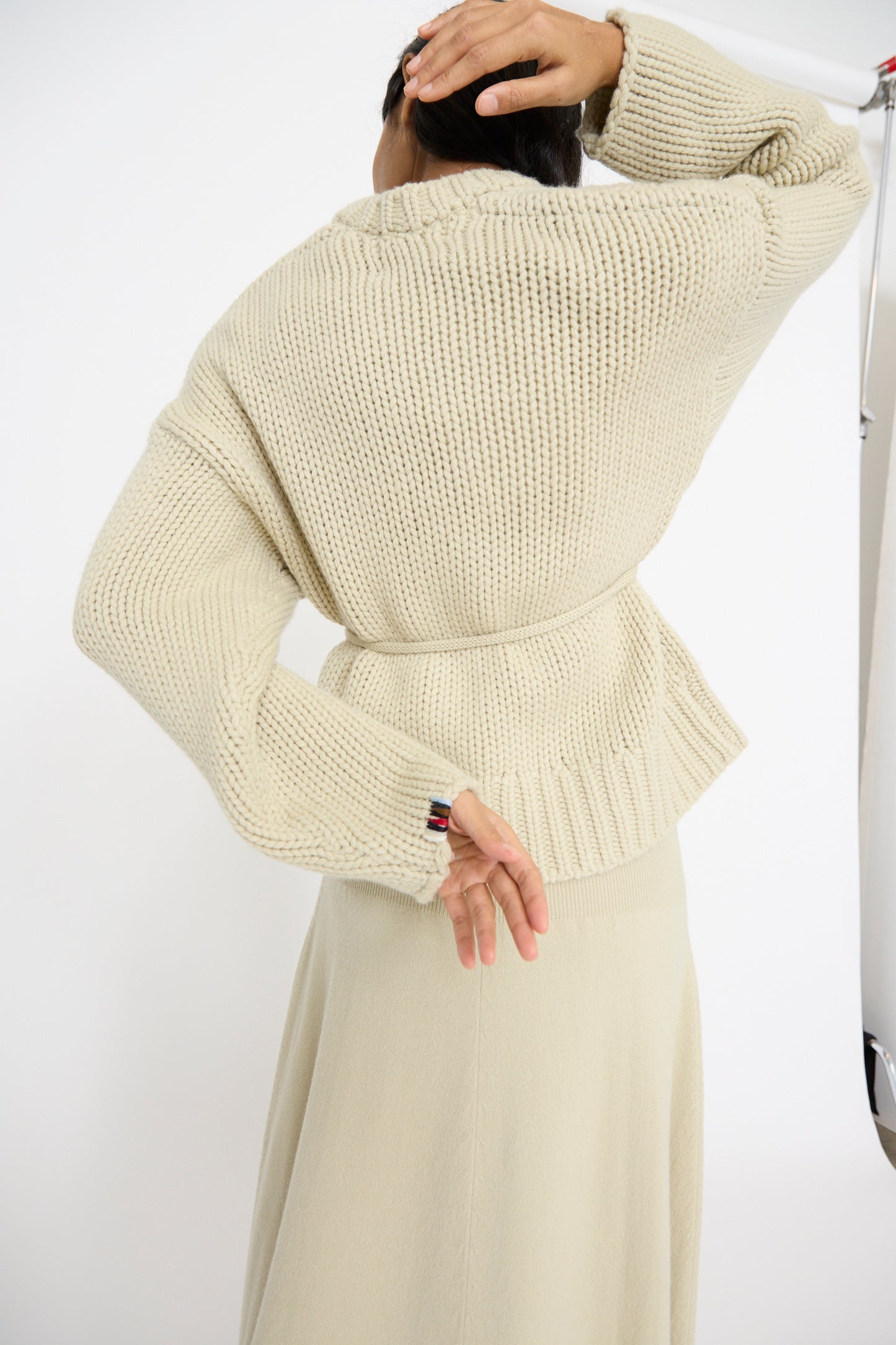 A person wearing an Extreme Cashmere No. 384 Ginza Handknit Cardigan in Desert adjusts their hair, facing away from the camera against a plain background.