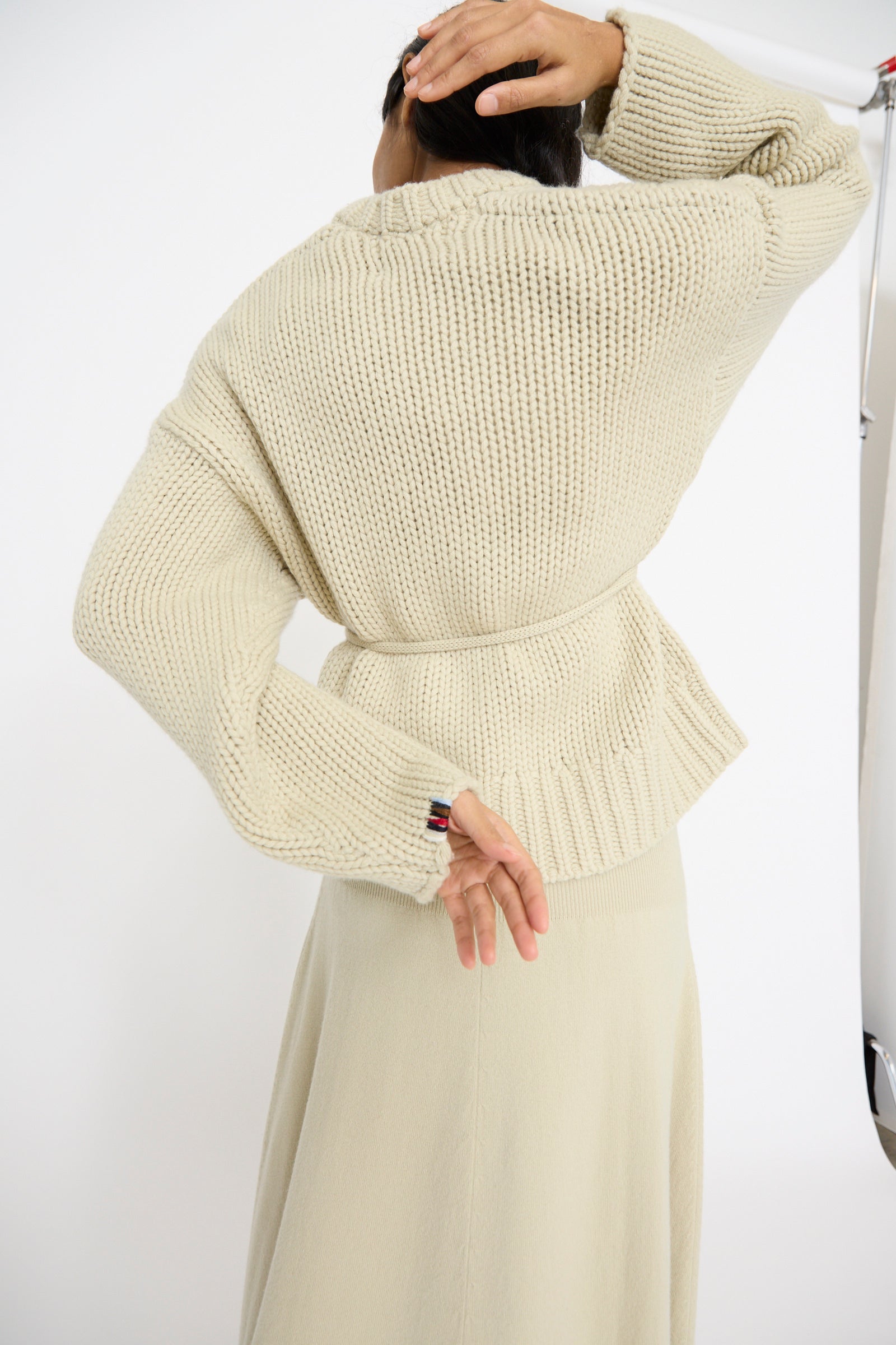 A person wearing an Extreme Cashmere No. 384 Ginza Handknit Cardigan in Desert adjusts their hair, facing away from the camera against a plain background.