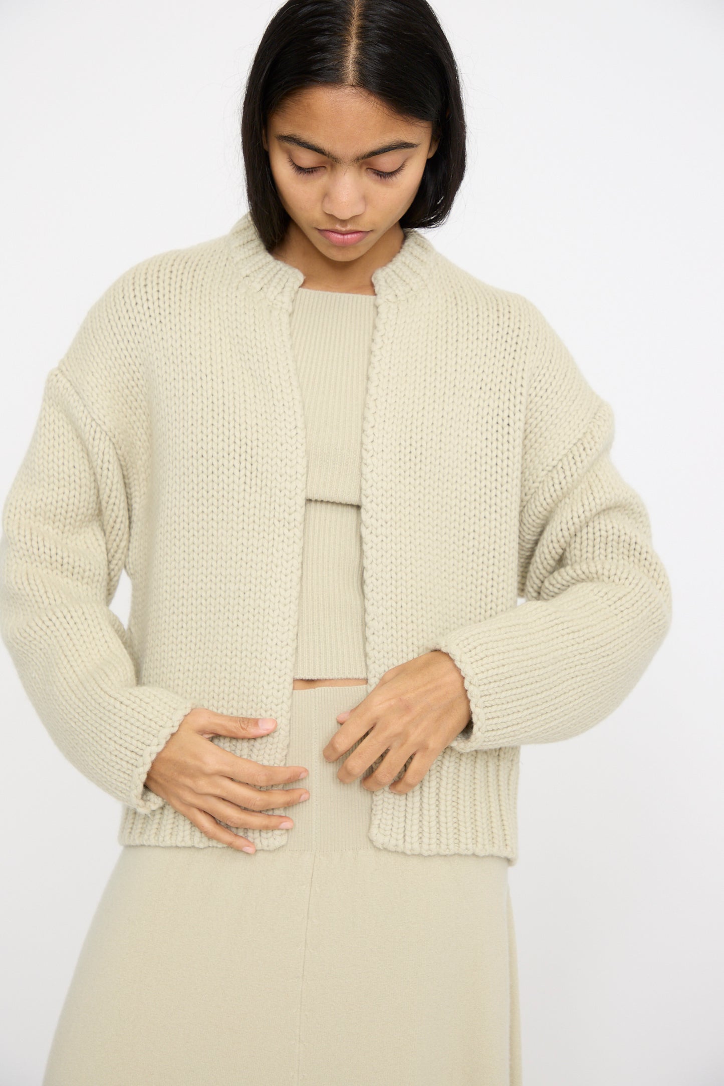 A woman is wearing the No. 384 Ginza Handknit Cardigan in Desert by Extreme Cashmere over a matching dress and looking down at her outfit against a white background.