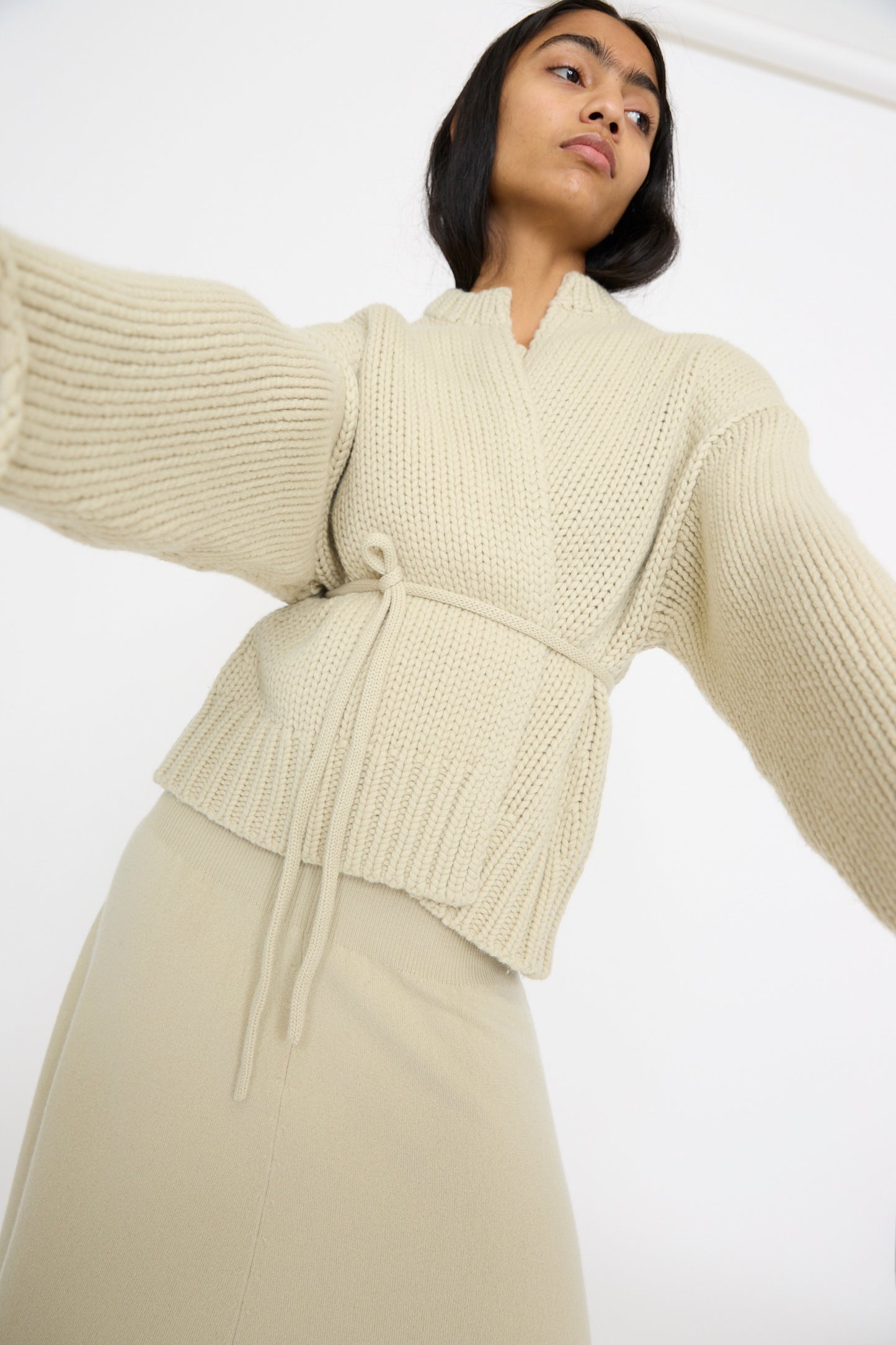 A person wearing the Extreme Cashmere No. 384 Ginza Handknit Cardigan in Desert with wide sleeves and a matching skirt poses gracefully against a white background.