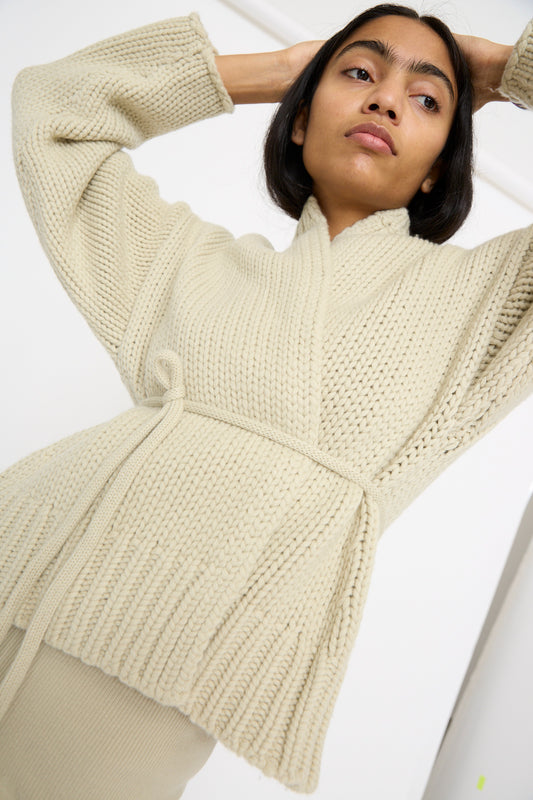 Person wearing the Extreme Cashmere No. 384 Ginza Handknit Cardigan in Desert, with a belt, arms raised and looking upward.