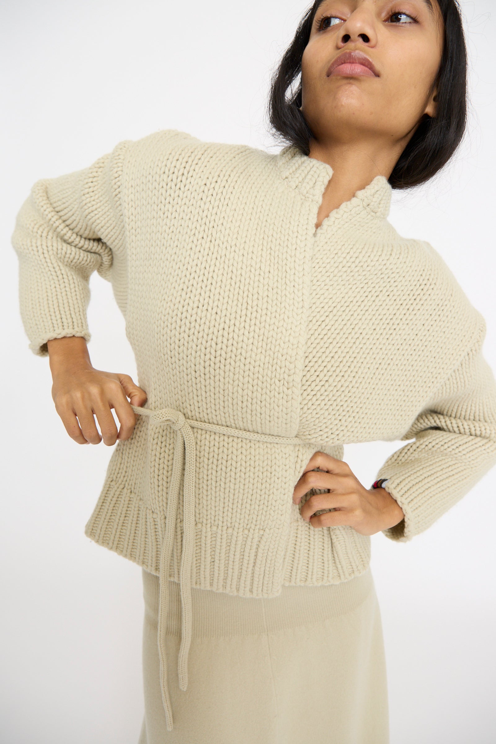 A person poses with hands on hips in a No. 384 Ginza Handknit Cardigan in Desert by Extreme Cashmere against a plain white background.