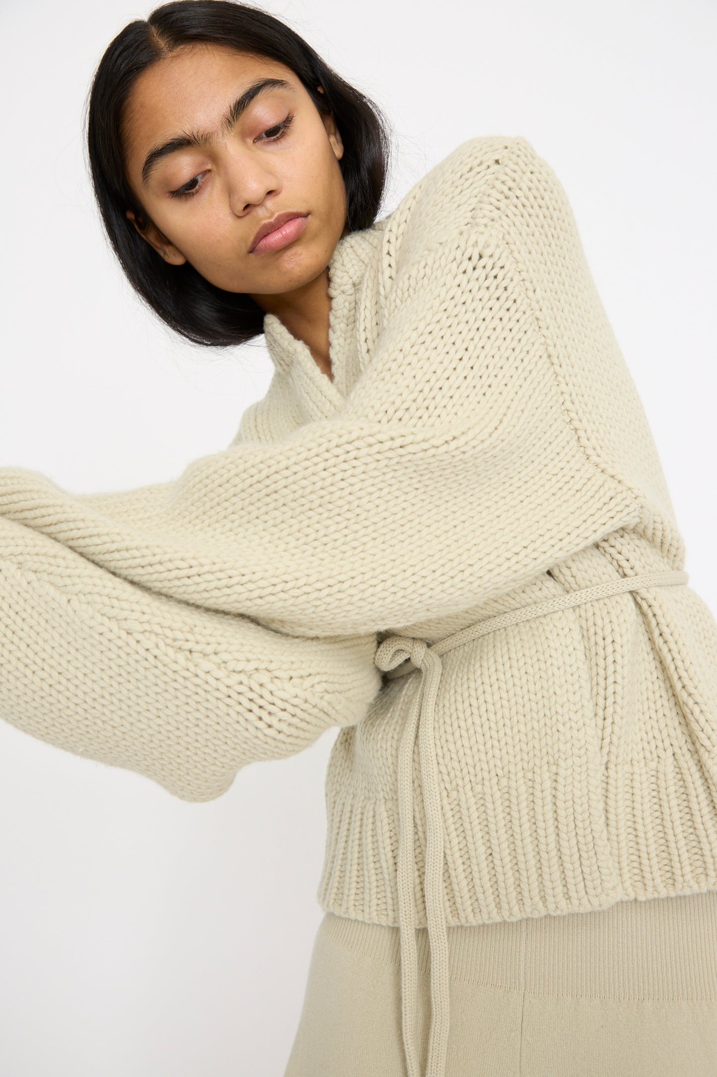 A person in the No. 384 Ginza Handknit Cardigan in Desert by Extreme Cashmere, gazes downward against a plain background.