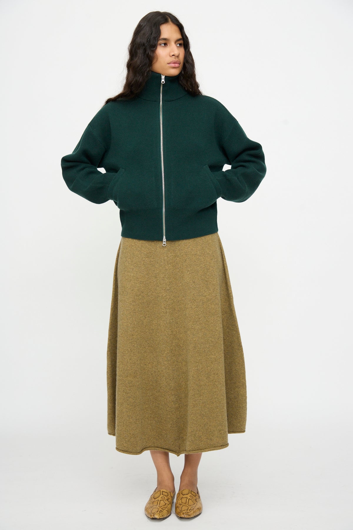 A person stands against a plain background wearing the Extreme Cashmere No. 385 Xtra Cute Cardigan in Forest, a long tan skirt, and tan patterned slip-on shoes.