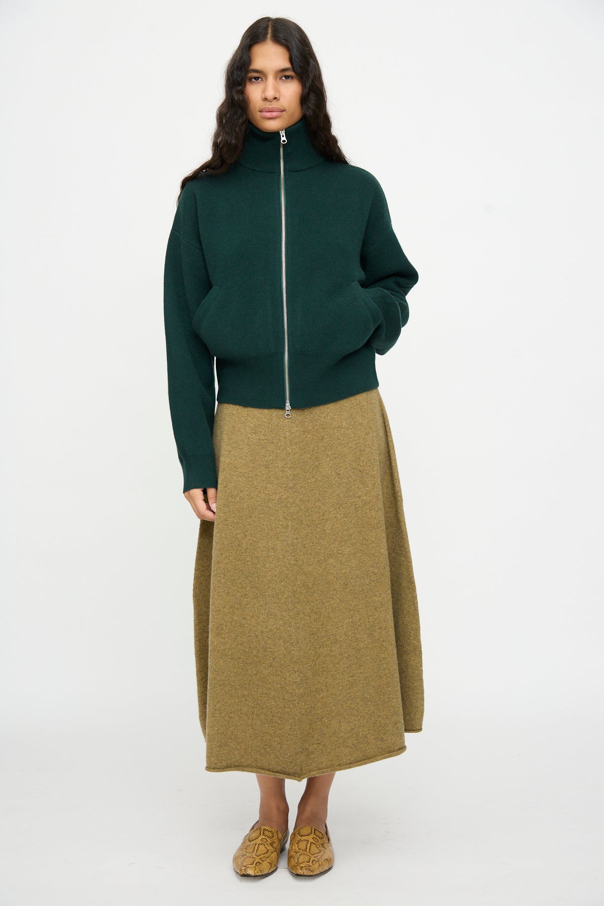 A woman stands facing forward against a plain white background, wearing the Extreme Cashmere No. 385 Xtra Cute Cardigan in Forest, an olive green midi skirt, and tan slip-on shoes.