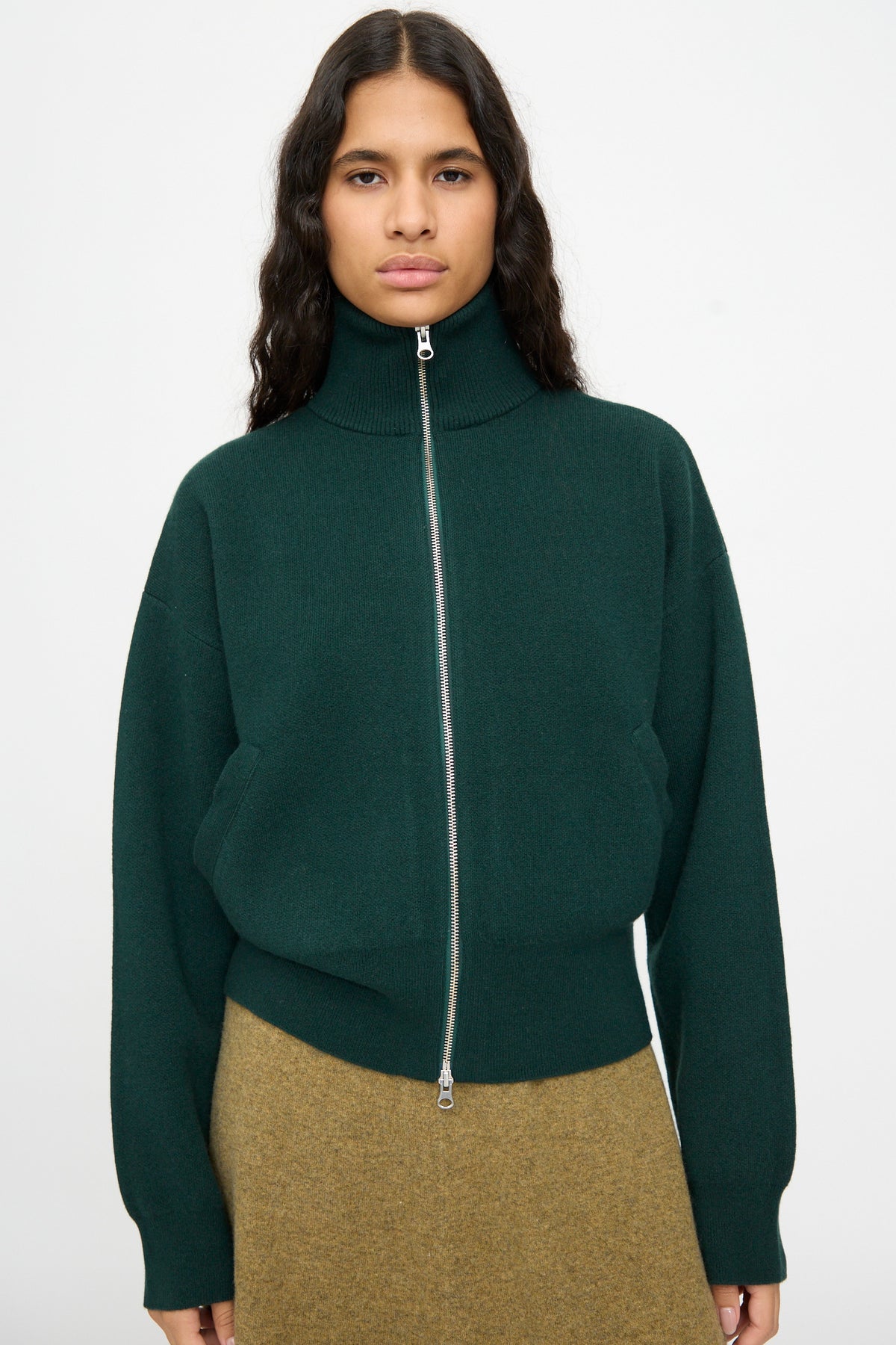 A person wearing the Extreme Cashmere No. 385 Xtra Cute Cardigan in Forest, styled with a brown skirt, stands against a plain light background.