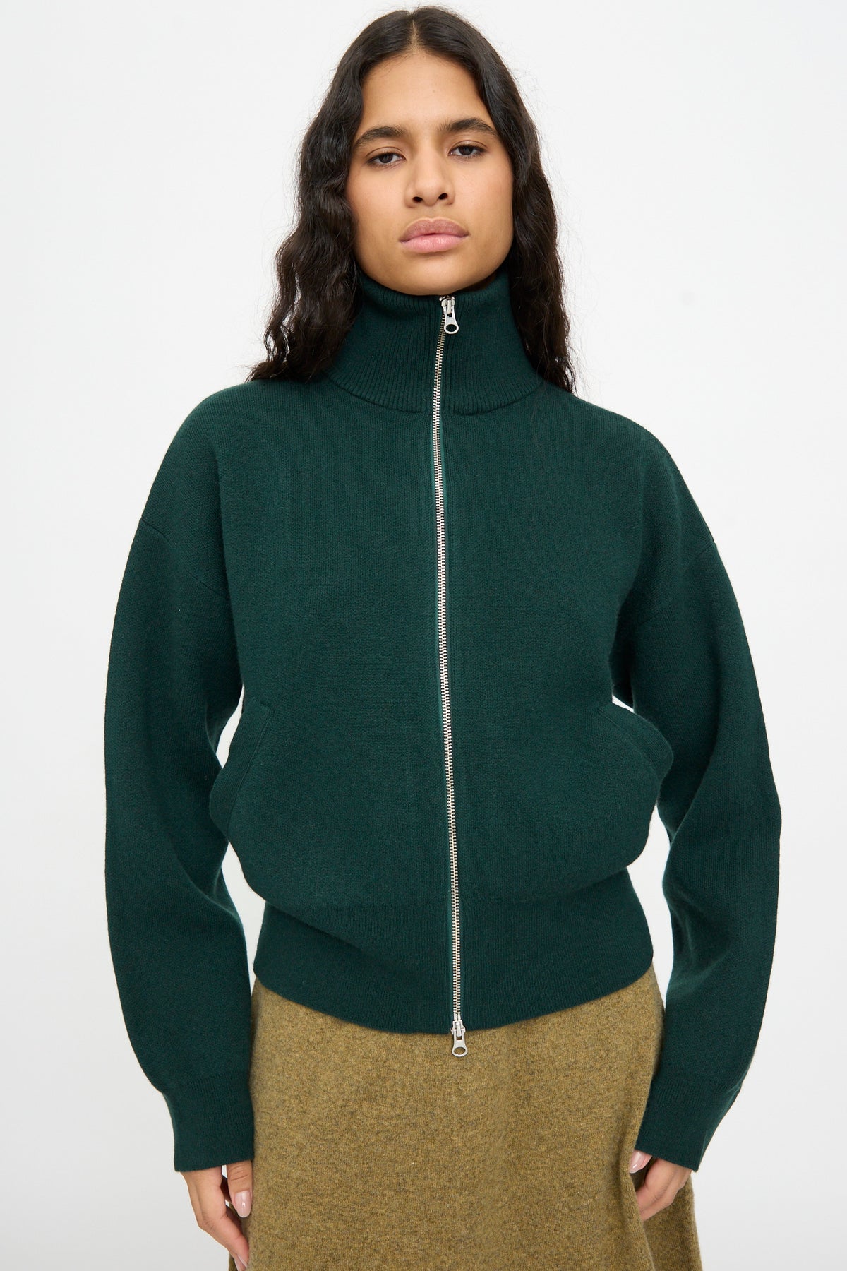 A person is wearing the Extreme Cashmere No. 385 Xtra Cute Cardigan in Forest, styled with a brown skirt and standing against a plain white background.