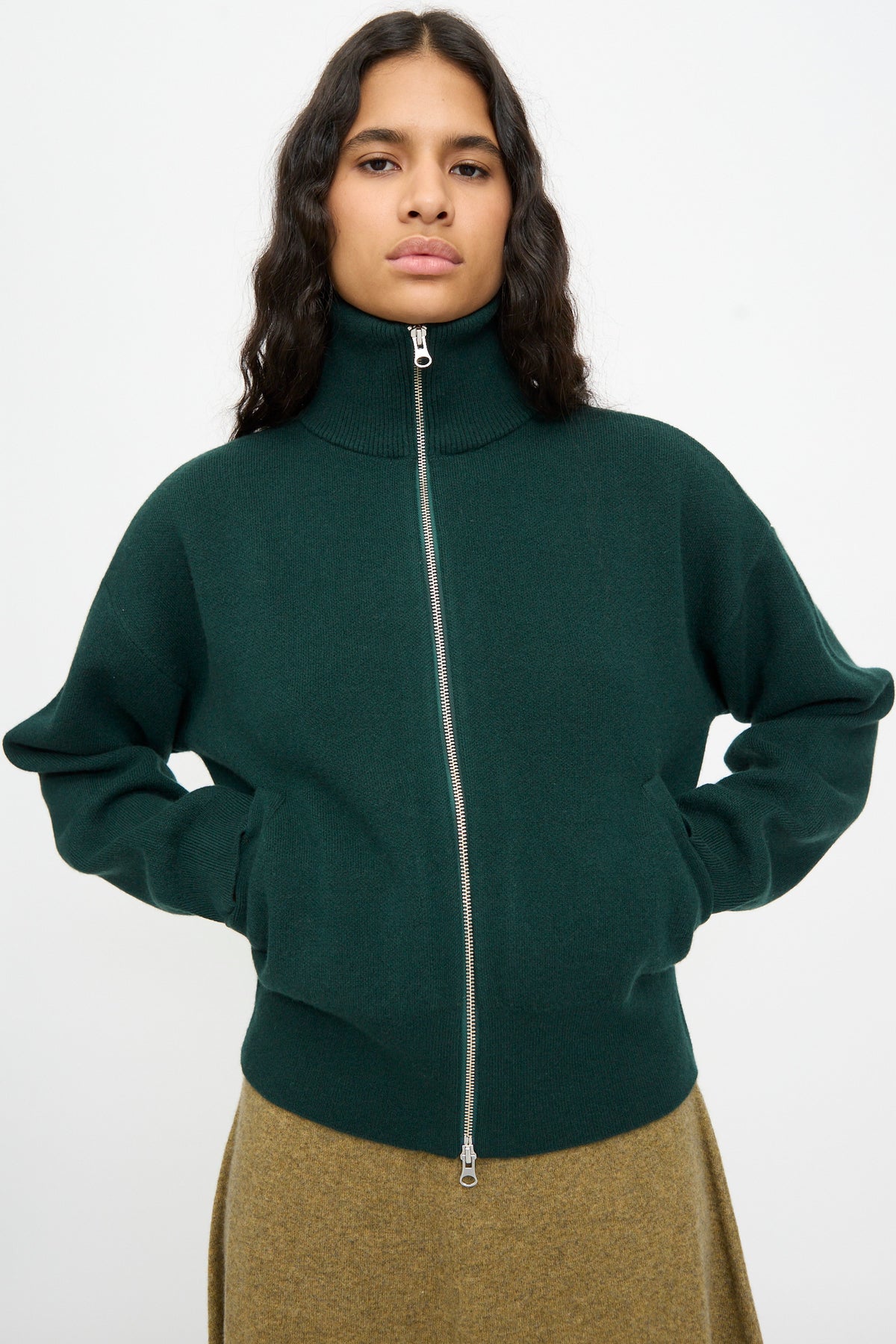 A person wearing the Extreme Cashmere No. 385 Xtra Cute Cardigan in Forest and a brown skirt, standing with hands in pockets against a plain light background.