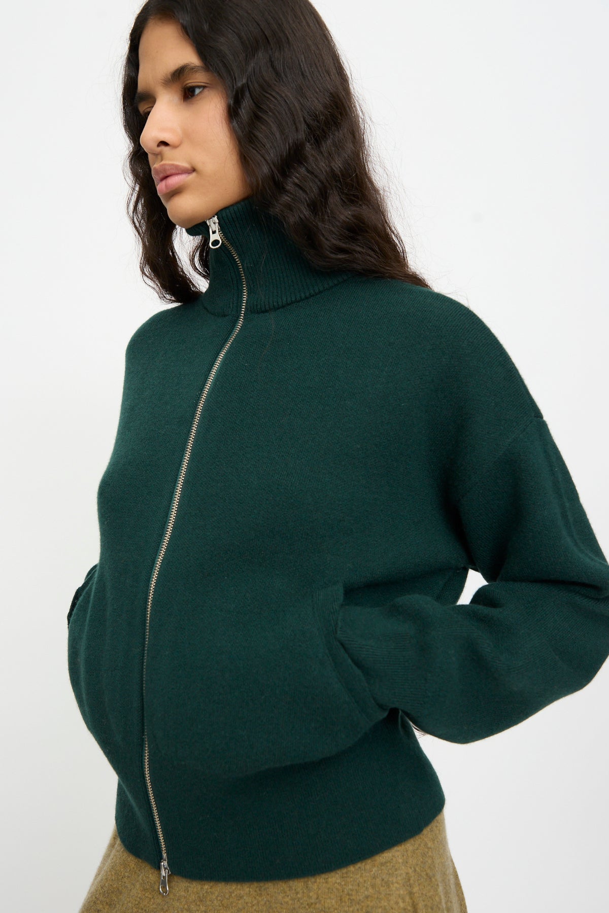 A person wearing the Extreme Cashmere No. 385 Xtra Cute Cardigan in Forest, hands in pockets, standing against a plain light background.