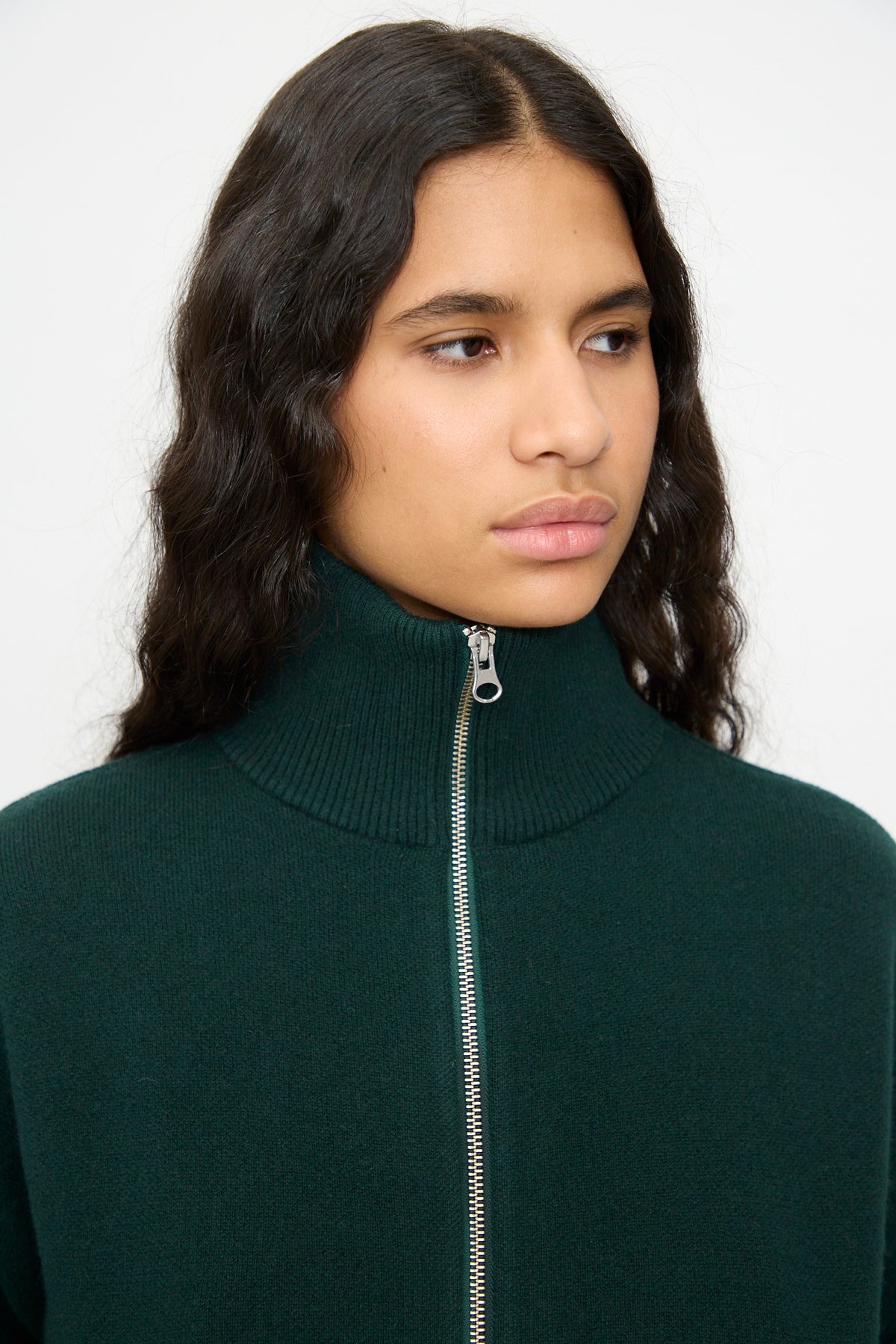 A person with long, wavy dark hair is wearing Extreme Cashmere’s No. 385 Xtra Cute Cardigan in Forest, layered over a dark green zip-up turtleneck sweater. They are facing slightly to the side with a neutral expression.