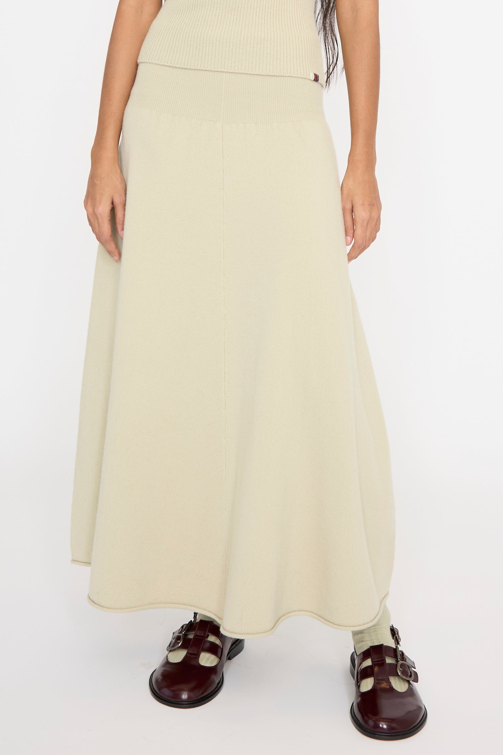 A person stands against a plain white background, wearing the No. 388 Ease Skirt in Desert by Extreme Cashmere, featuring a rib-knitted waistline, paired with brown shoes, seen from below the waist. 