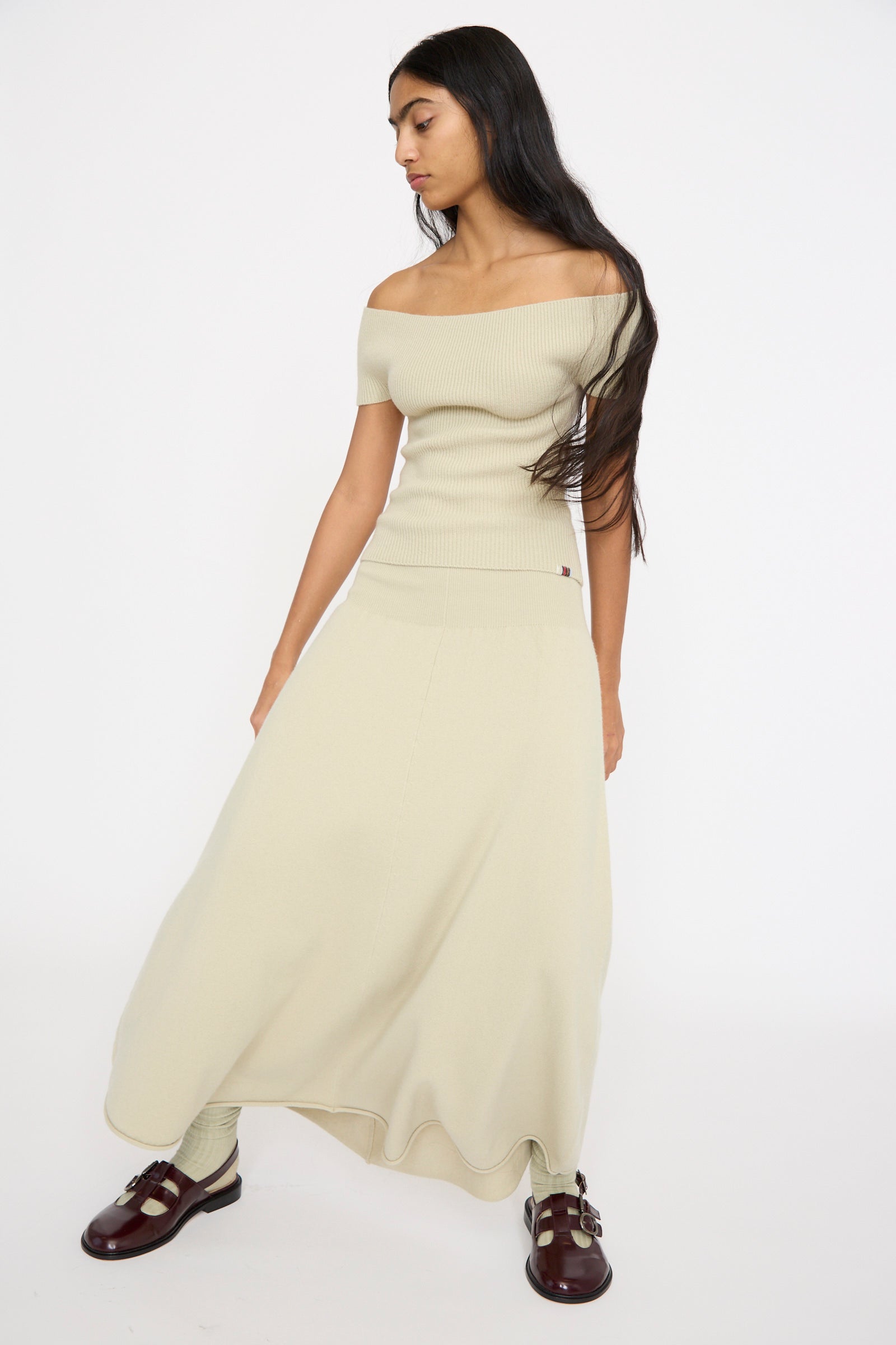 A person stands against a plain background, looking downward. They have long dark hair and are wearing red shoes, paired with an off-shoulder beige top and the Extreme Cashmere No. 388 Ease Skirt in Desert, featuring a flared design and rib-knitted waistline.