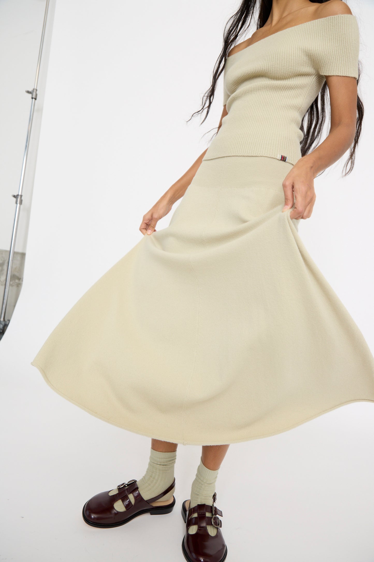 A person in a beige off-shoulder top and Extreme Cashmere's No. 388 Ease Skirt in Desert with a rib-knitted waist stands against a white background, with a pair of dark brown shoes. 