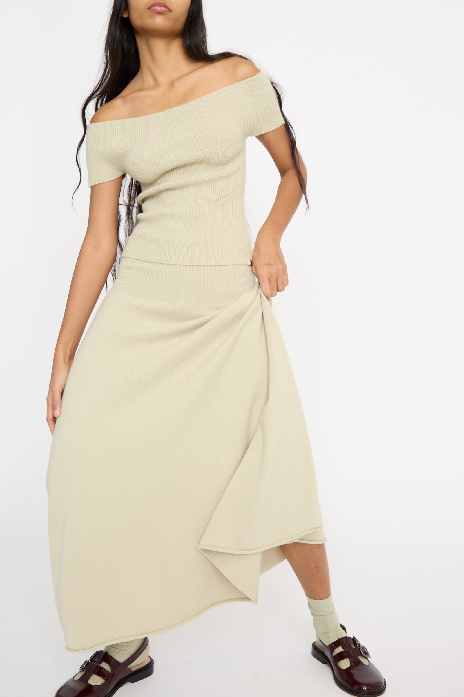 A person in an off-the-shoulder beige top and the No. 388 Ease Skirt in Desert by Extreme Cashmere, featuring a flared silhouette with a rolling edge hem, stands with long hair over one shoulder against a plain background.