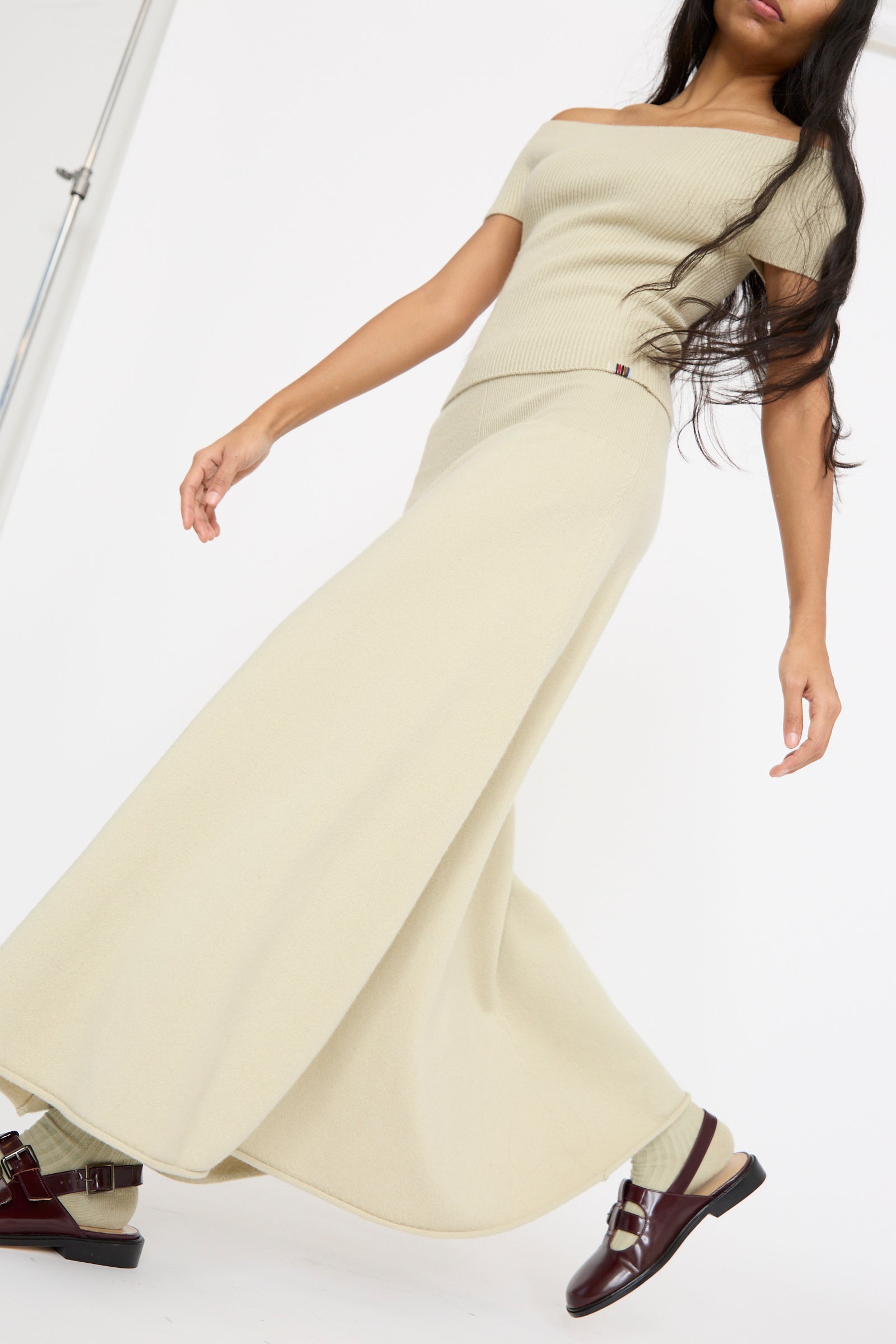 Against a white backdrop, an individual models the No. 388 Ease Skirt in Desert by Extreme Cashmere, a light beige ensemble with a ribbed top and matching long skirt featuring a rib-knitted waistline. 