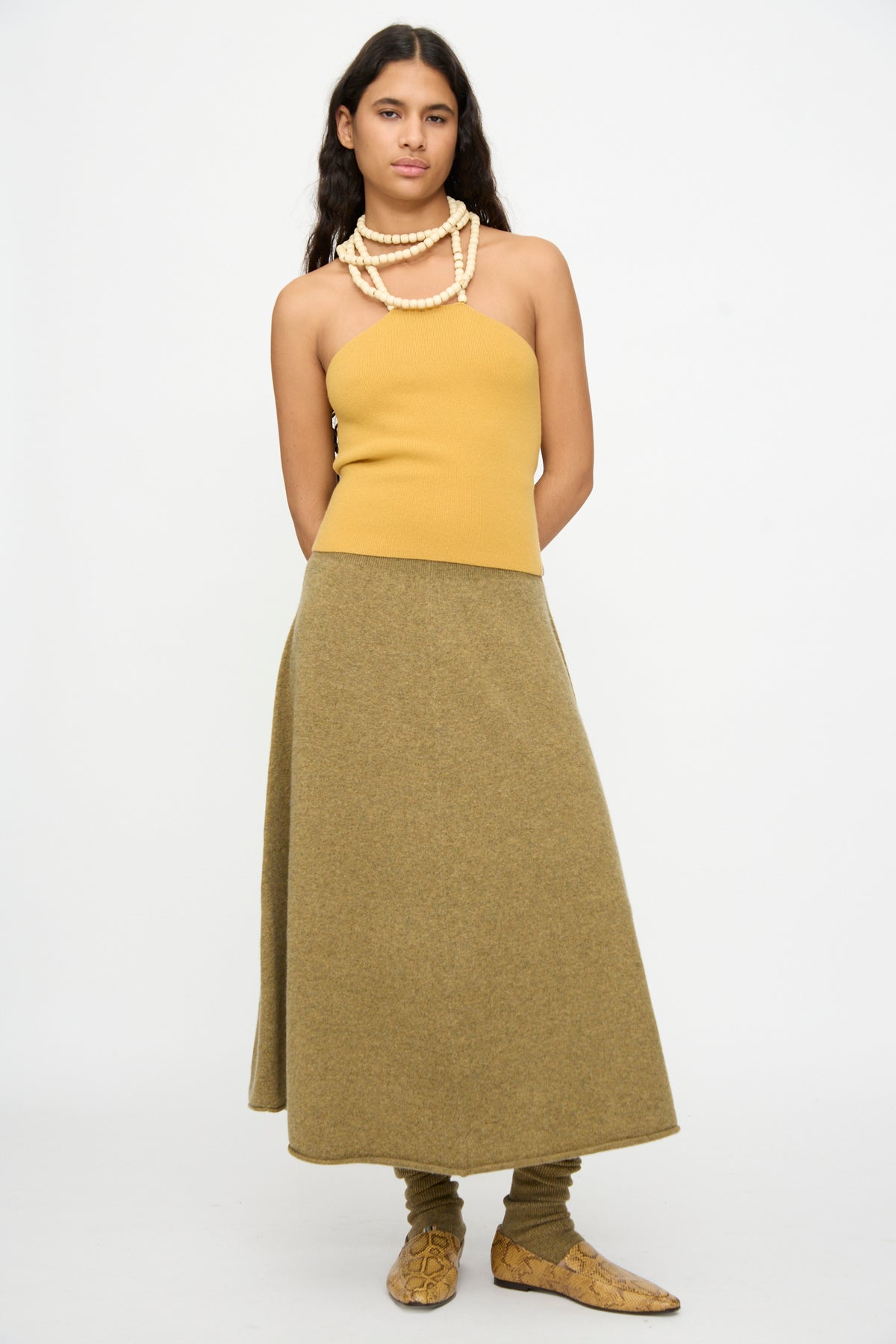 A person stands against a plain background wearing a yellow strapless top, the Extreme Cashmere No. 388 Ease Skirt in Hunter—a flared olive green skirt with a rib-knitted waistline—paired with matching leg warmers, beige shoes, and layered beaded necklaces.