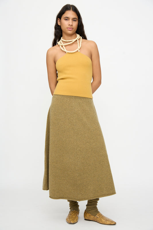 A person stands against a plain background wearing a yellow strapless top, the Extreme Cashmere No. 388 Ease Skirt in Hunter—a flared olive green skirt with a rib-knitted waistline—paired with matching leg warmers, beige shoes, and layered beaded necklaces.