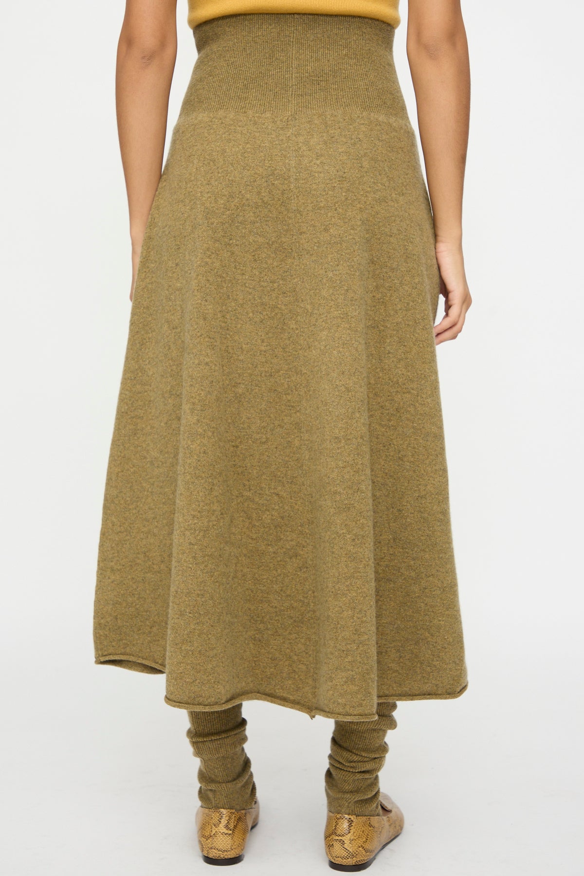 A person stands facing away, wearing the Extreme Cashmere No. 388 Ease Skirt in Hunter—an ankle-length, high-waisted olive-green flared skirt with a rib-knitted waistline—paired with matching leg warmers and snakeskin-patterned shoes, set against a plain white background.