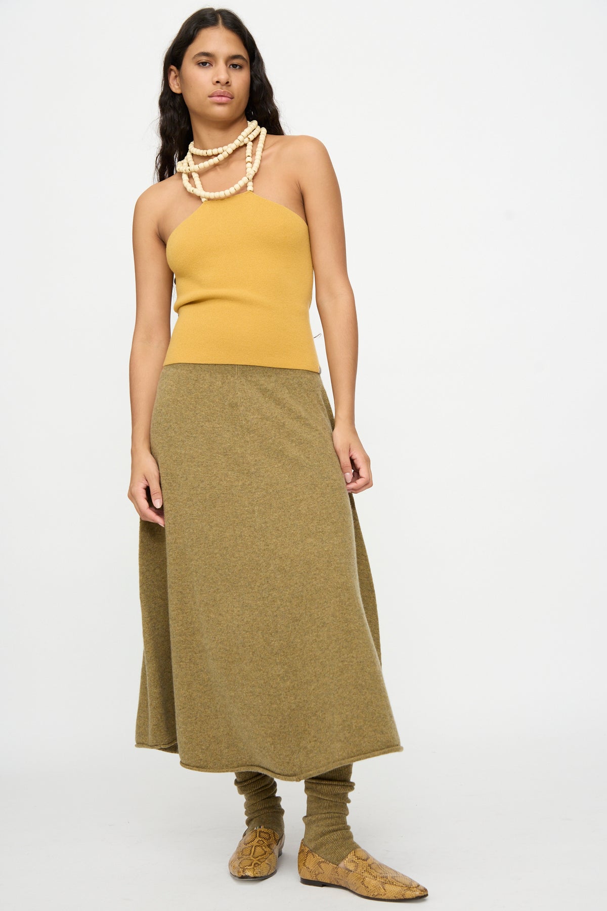 A woman stands against a plain white background wearing Extreme Cashmere’s No. 388 Ease Skirt in Hunter—an olive green flared skirt with a rib-knitted waistline—paired with a mustard yellow one-shoulder top, layered necklaces, and tan textured shoes.