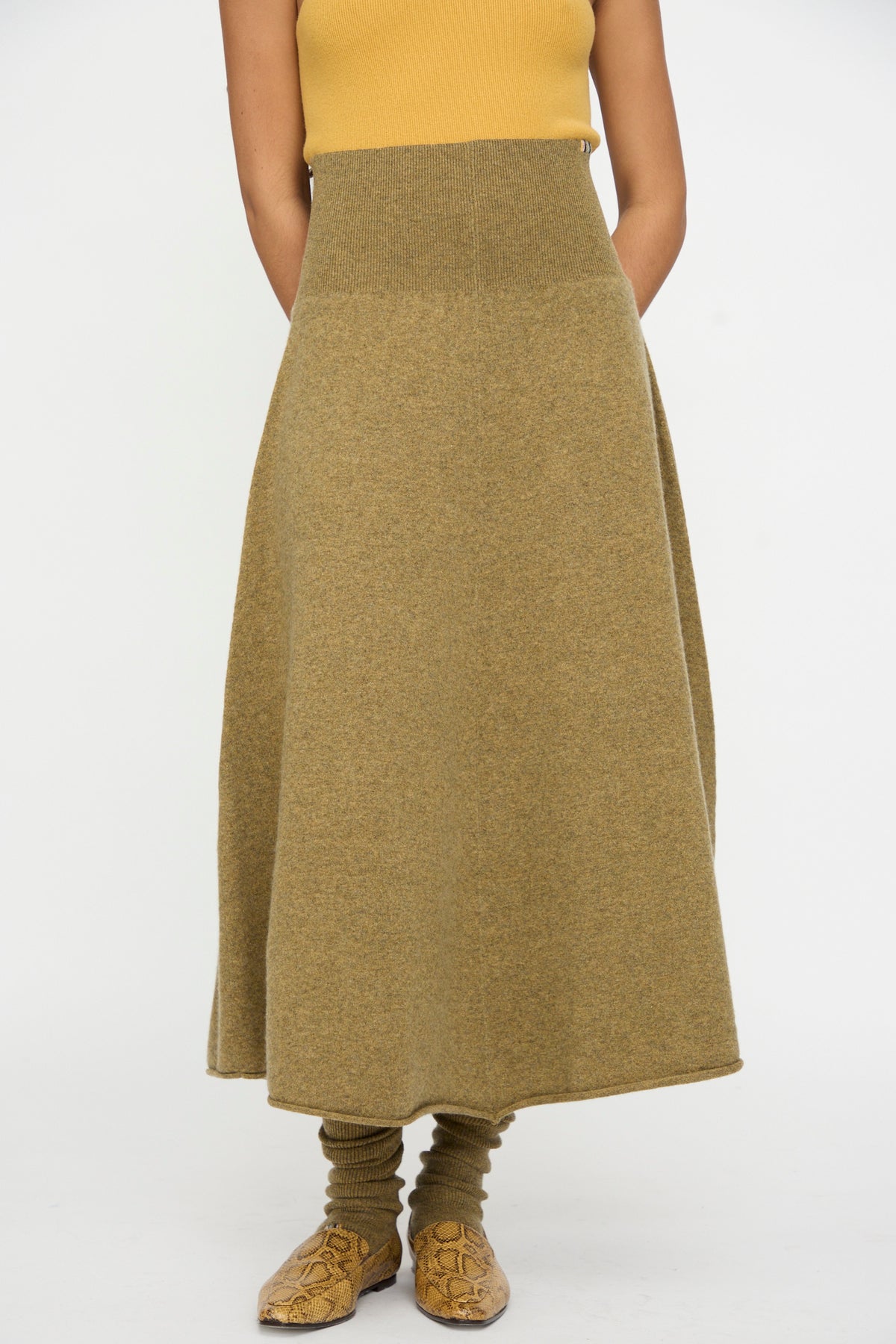 A person is wearing a mustard sleeveless top, the No. 388 Ease Skirt in Hunter by Extreme Cashmere—a high-waisted olive cashmere midi skirt with a rib-knitted waistline—paired with matching leg warmers and tan patterned shoes, standing against a plain white background.