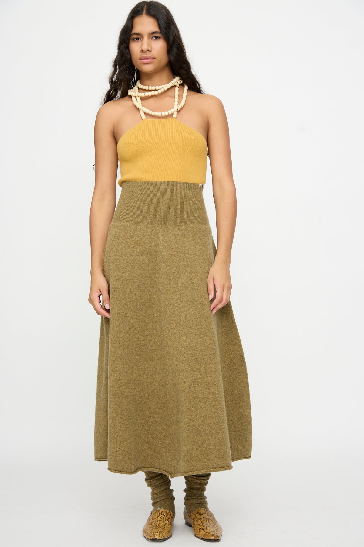 A woman stands against a plain background, wearing a mustard yellow strapless top, the Extreme Cashmere No. 388 Ease Skirt in Hunter—an olive green cashmere midi skirt with a rib-knitted waistline—matching leg warmers, brown shoes, and layered beaded necklaces.
