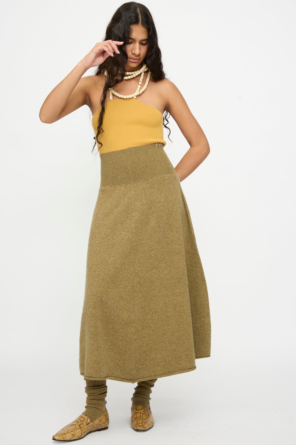 Woman wearing a mustard strapless top, the Extreme Cashmere No. 388 Ease Skirt in Hunter—an olive green high-waisted cashmere midi skirt with a rib-knitted waistline—matching leg warmers, tan boots, and layered pearl necklaces, standing against a plain white background.