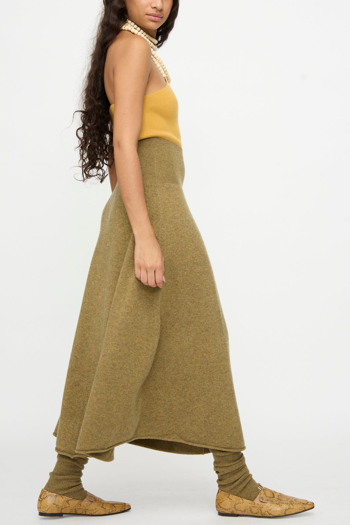 A woman in a yellow top and the Extreme Cashmere No. 388 Ease Skirt in Hunter, featuring a rib-knitted waistline, pairs the look with beige snakeskin shoes and layered pearl necklaces as she walks against a plain white background.