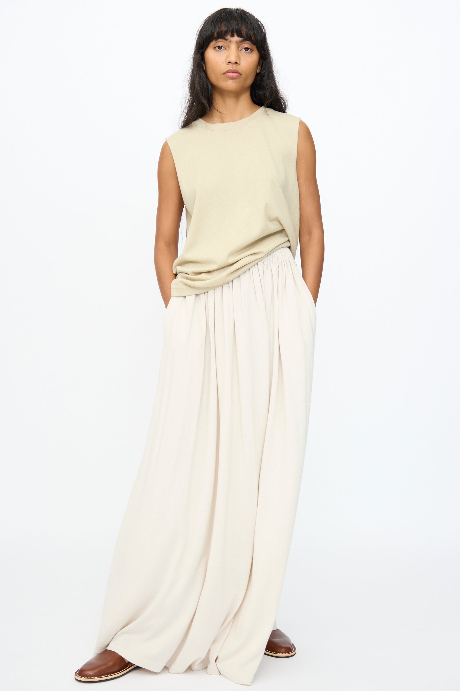 A person stands against a plain background wearing a sleeveless beige top, brown shoes, and the No. 403 Mary Skirt in Chalk by Extreme Cashmere—an extra long, relaxed fit cream cotton-cashmere skirt—with hands in their pockets.