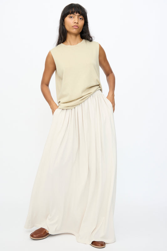 A woman stands against a plain backdrop wearing a sleeveless beige top, brown slip-on shoes, and the No. 403 Mary Skirt in Chalk by Extreme Cashmere.