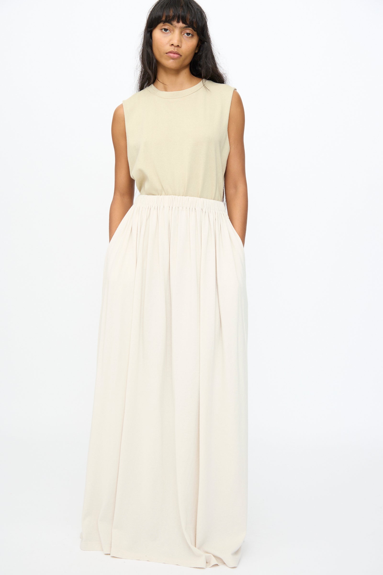 A woman stands against a plain background wearing a sleeveless beige top and the No. 403 Mary Skirt in Chalk by Extreme Cashmere—an extra-long, high-waisted cream skirt with pockets.