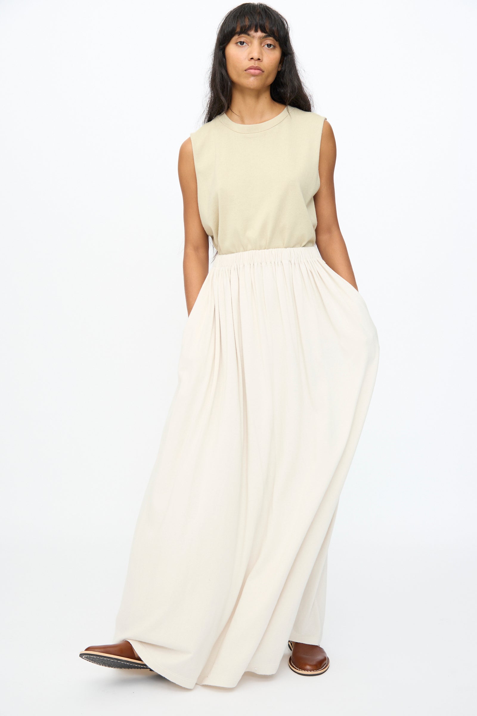 A person stands against a white background wearing a sleeveless beige top, brown shoes, and the Extreme Cashmere No. 403 Mary Skirt in Chalk—an extra-long, off-white relaxed fit skirt with pockets.