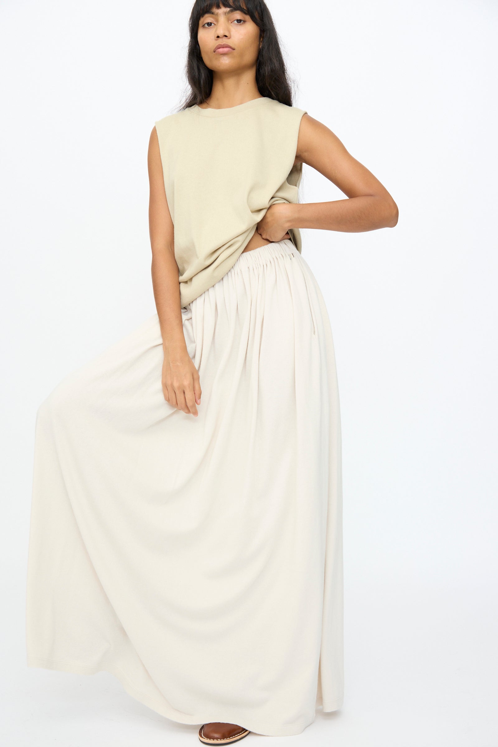 A woman poses wearing the No. 403 Mary Skirt in Chalk by Extreme Cashmere, paired with a sleeveless beige top, one hand on her hip and the other holding the flowing skirt.