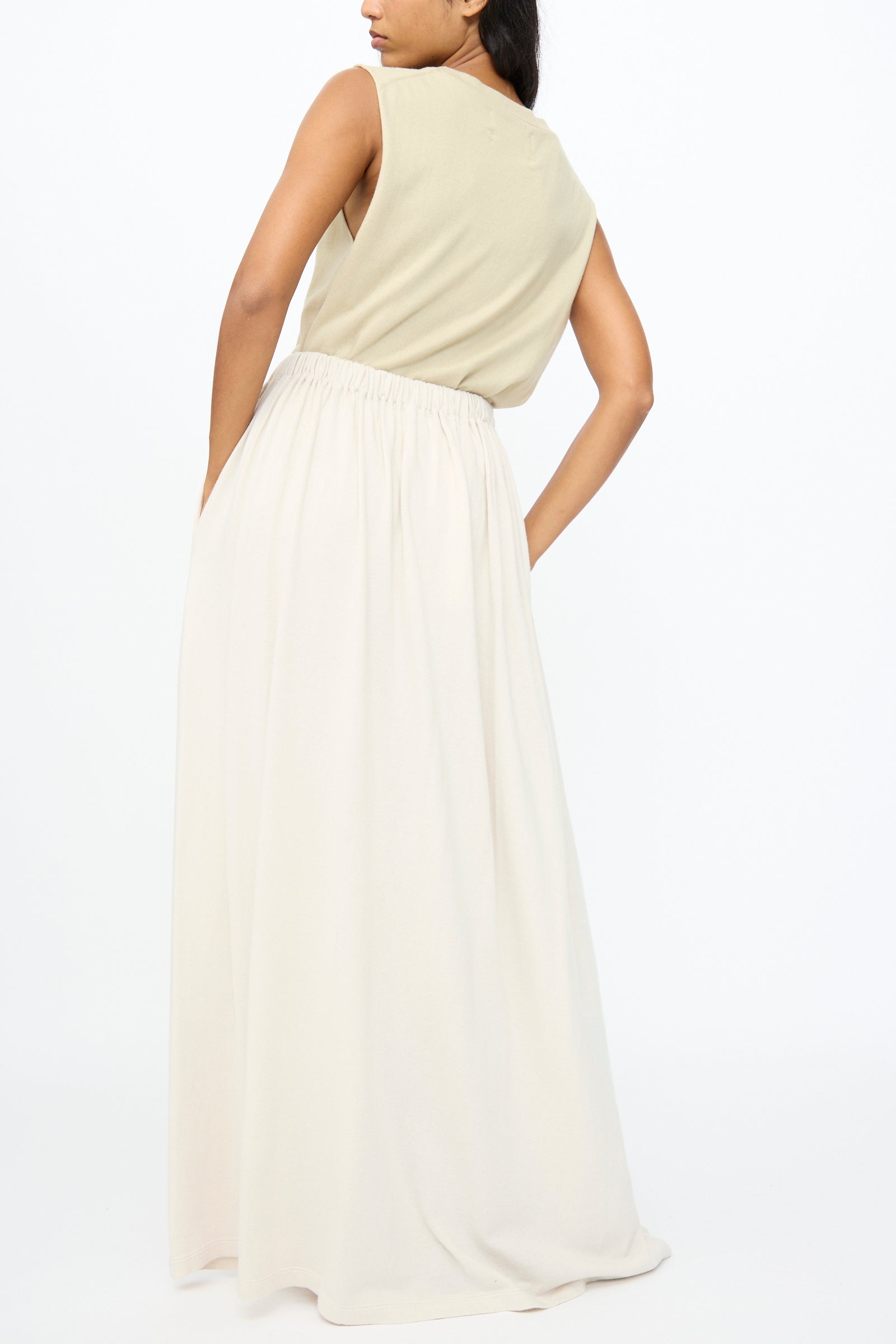 A person stands against a plain white background wearing Extreme Cashmere's No. 403 Mary Skirt in Chalk, styled with a sleeveless beige top and one hand behind their back.