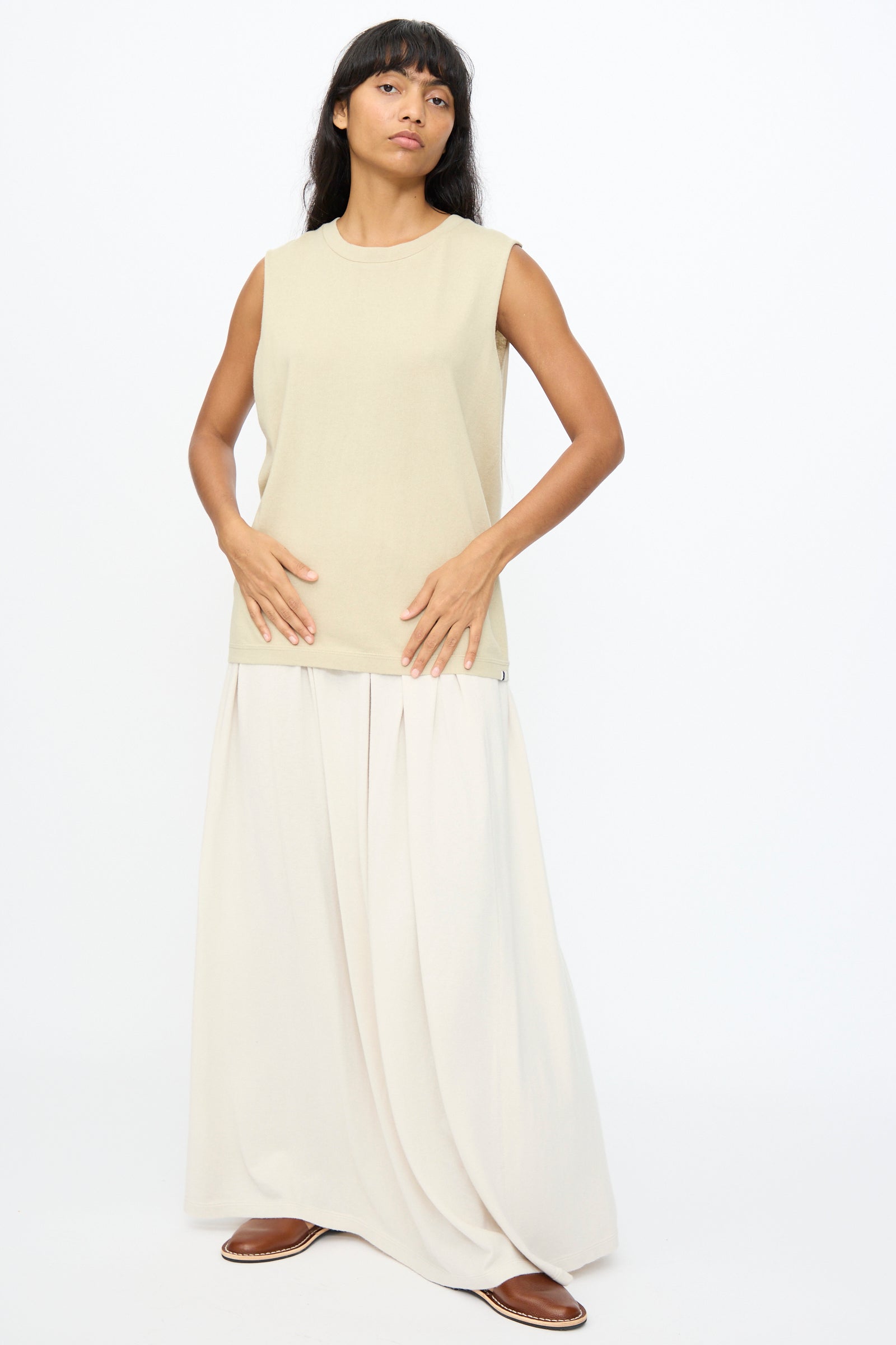 A woman stands against a plain white background wearing Extreme Cashmere's No. 408 Muscle Tee in Desert, paired with a long cream skirt and brown shoes.